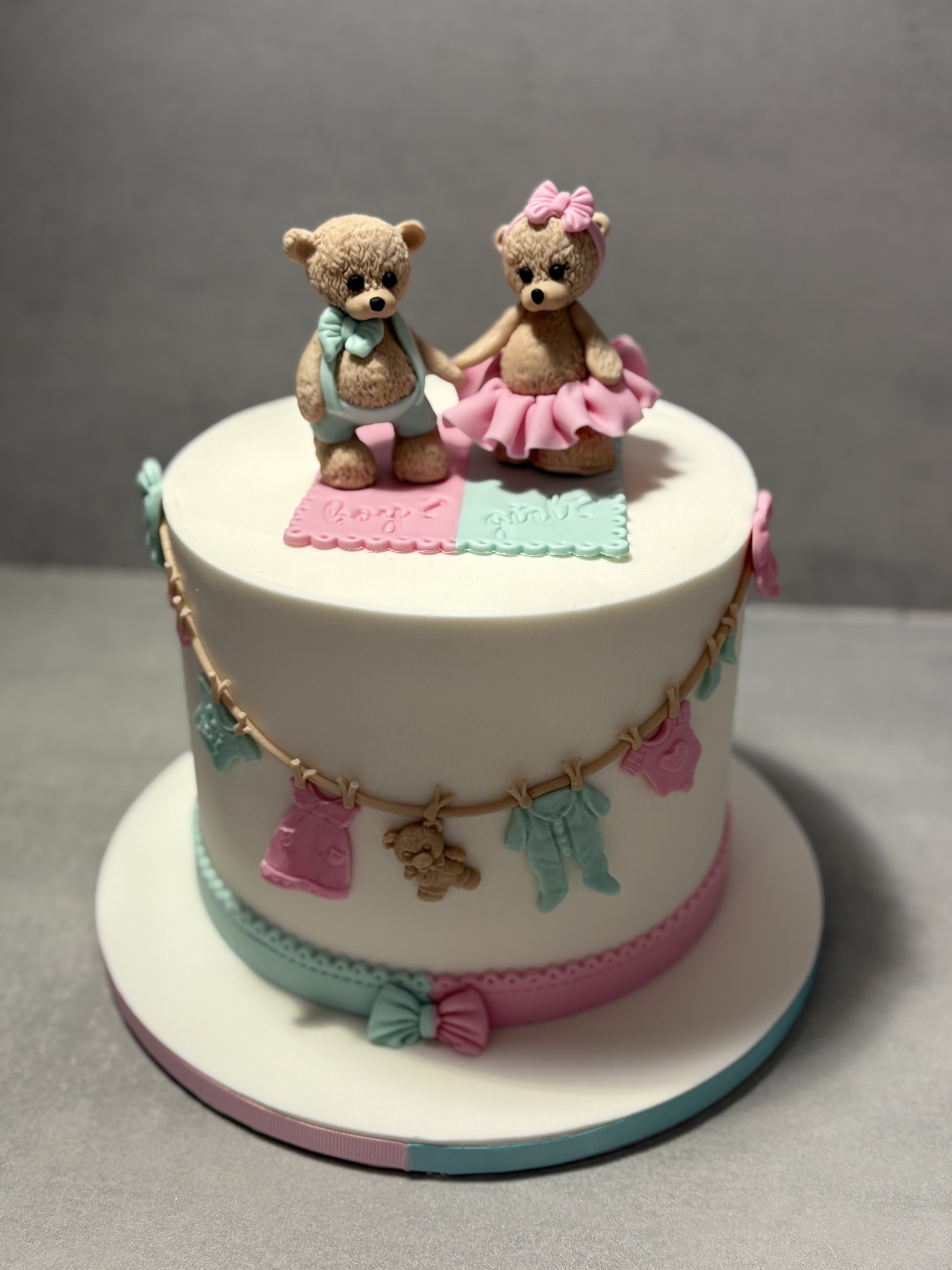 Gâteau blanc décoré avec deux ours en peluche minutieusement modelés en sucre, un homme et une femme se tenant la main, avec des détails en rose et en bleu, et une guirlande de habits de bébé en sucre autour du gâteau.