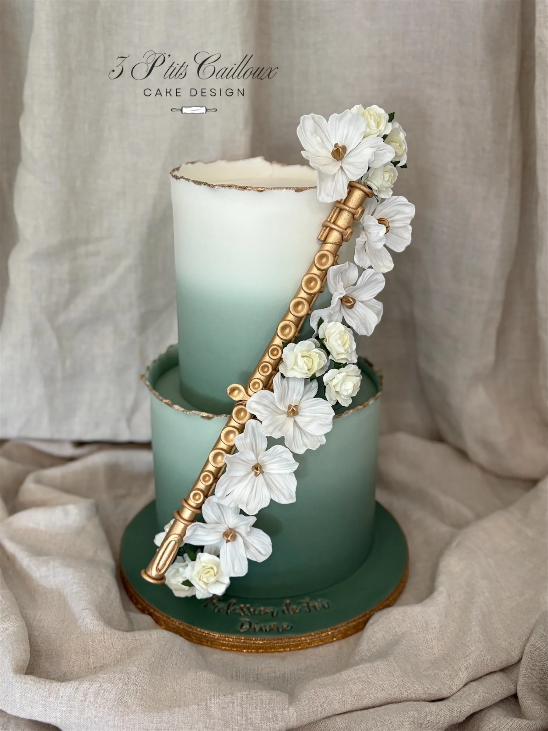 Un gâteau à deux niveaux avec une décoration florale blanche, un flûte en or, et un fond en tissu beige.