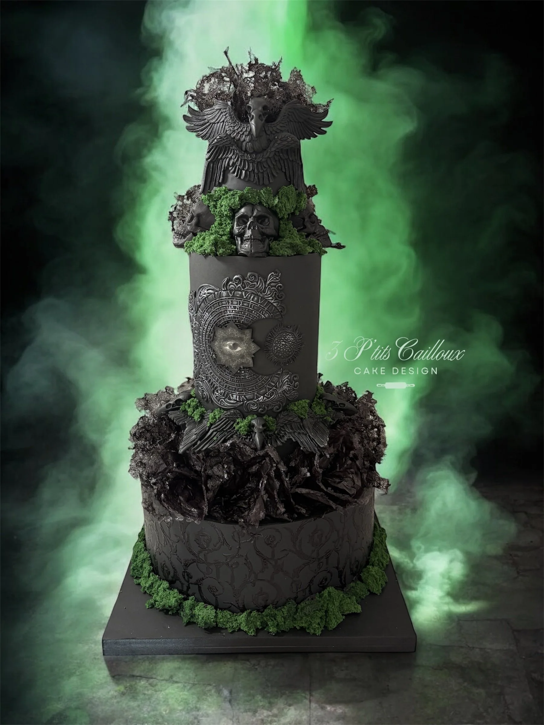 Gâteau noir à plusieurs étages avec des décorations mystiques, comprenant un aigle en haut, un crâne, des symboles et des matières organiques, entouré de fumée verte.