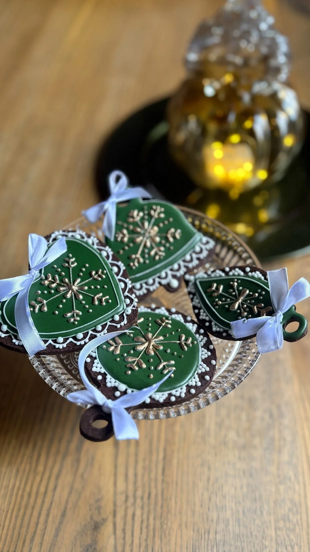 Sablés de Noël décorés de glaçage vert, ornement en forme de flocon de neige avec des détails blancs et dorés, liés par des rubans blancs, disposés sur un plat en verre