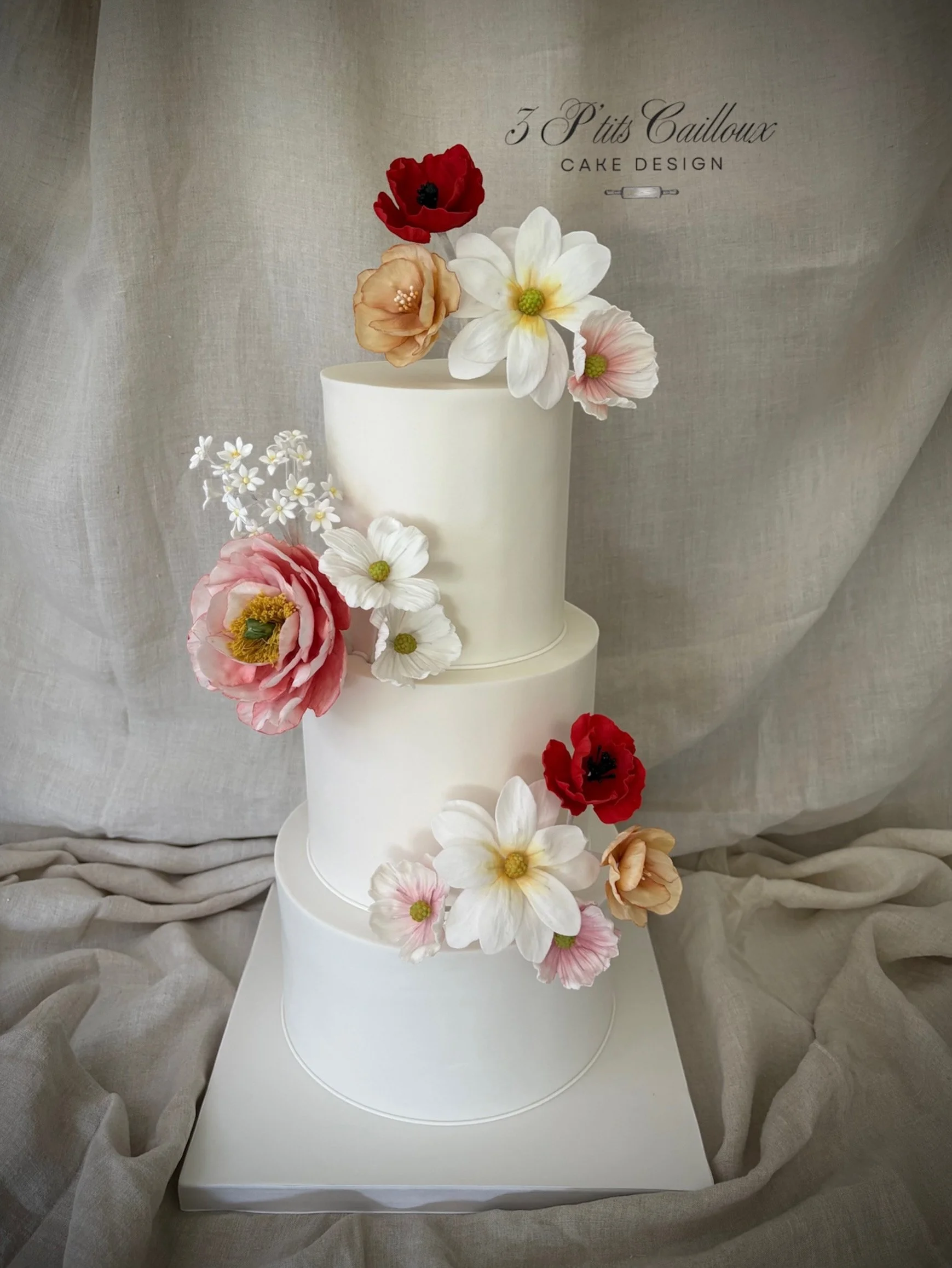 Gâteau blanc à trois niveaux décoré de fleurs en artificiel de différentes couleurs, avec un fond en tissu léger.