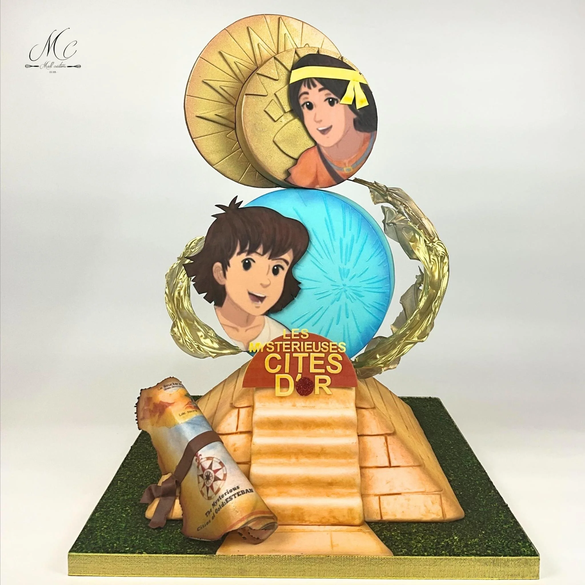 Une décoration représentant deux personnages avec des visages illustrés, inspirés de dessins animés, sur une base en forme de pyramide. Il y a aussi un rouleau de parchemin attaché avec un ruban, et un panneau indiquant « Les mystérieuses cîtés d'or 