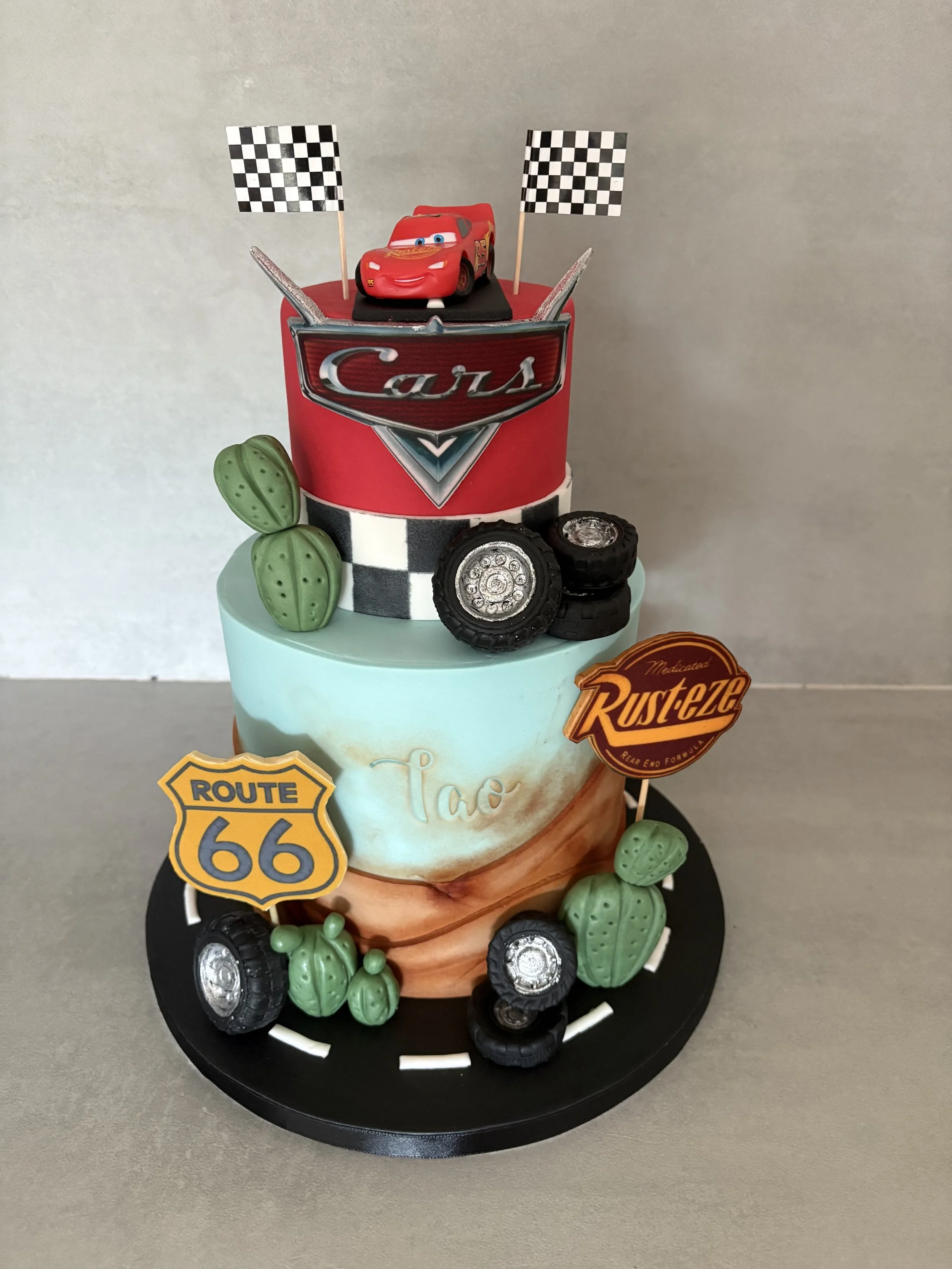 Gâteau à plusieurs niveaux décoré avec des motifs de voitures, pneus, cactus, et symboles liés à la course, avec une figurine de voiture de Pixar sur le dessus, et des drapeaux de course.
