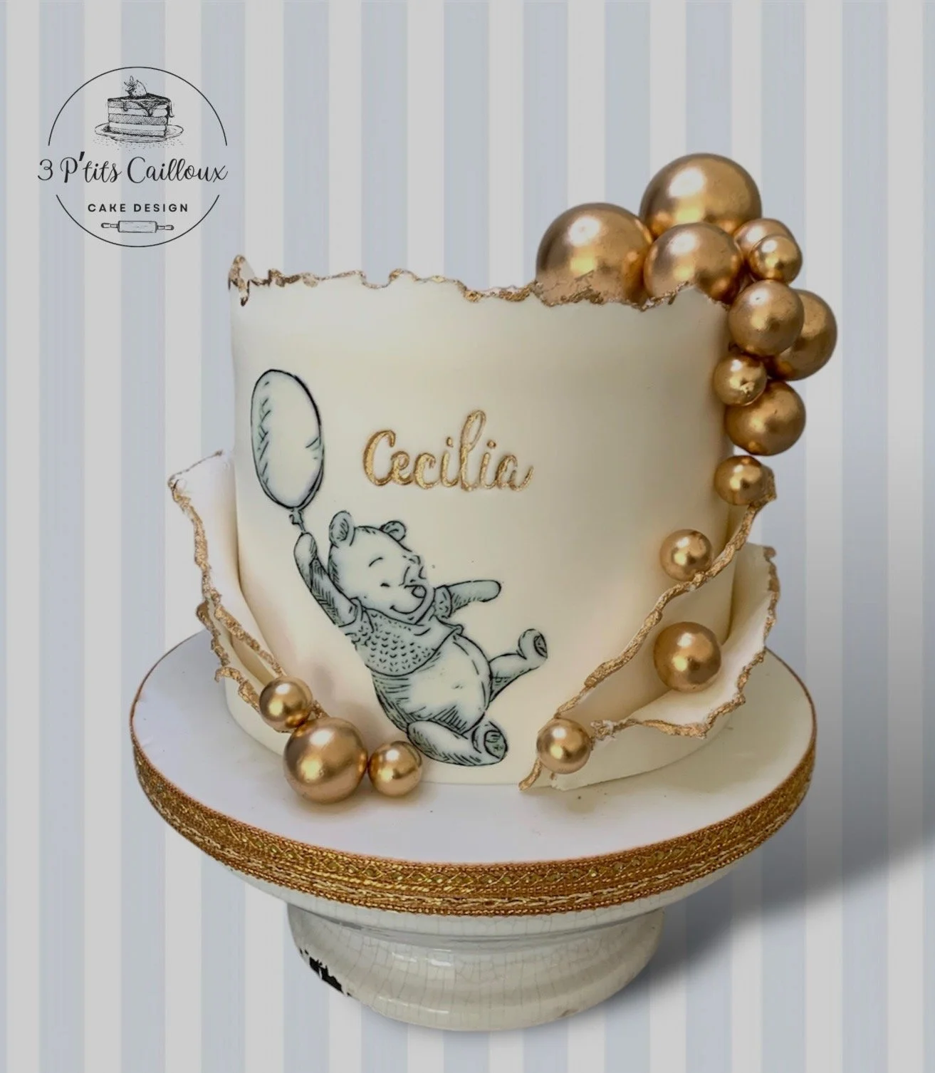 Gâteau blanc décoré de perles dorées, avec une illustration de Winnie l'Ourson tenant un ballon et le nom 'Cecilia' écrit en or, posé sur une piédestal blanc avec détails dorés.