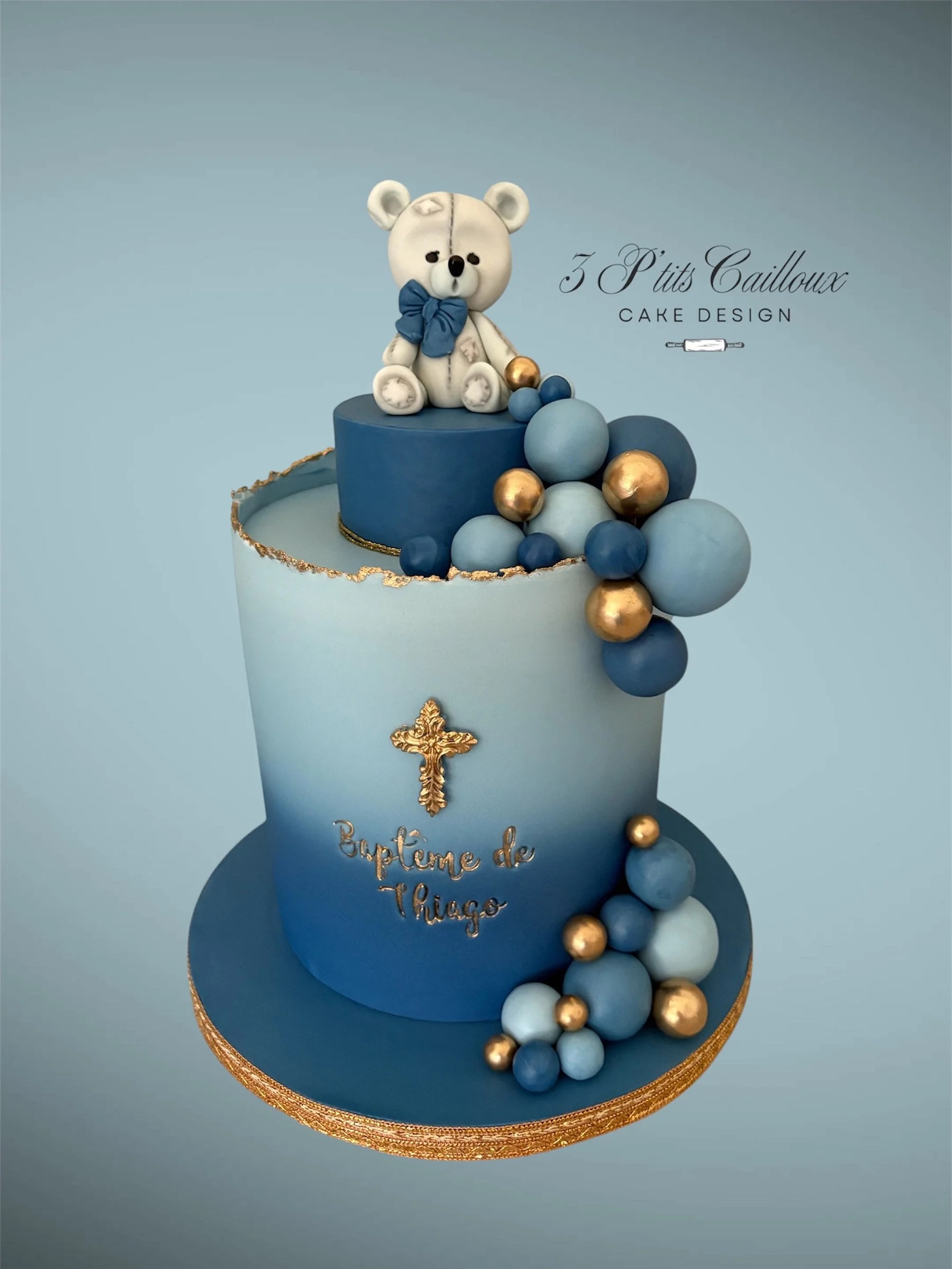 Gâteau d'inspiration religieuse ou religieuse avec un ours en peluche blanc portant un nœud bleu sur le dessus, décoré de boules de différentes tailles en bleu, blanc et or, avec une croix dorée et le texte 'Baptême de Thiago'.