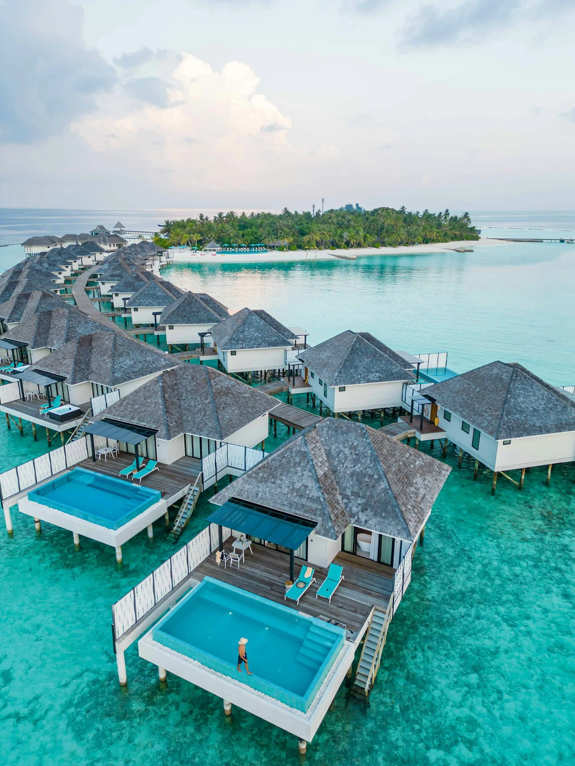 Overwater bungalows with private pools and decks in clear turquoise water near a tropical island with palm trees and a sandy beach, under a cloudy sky.