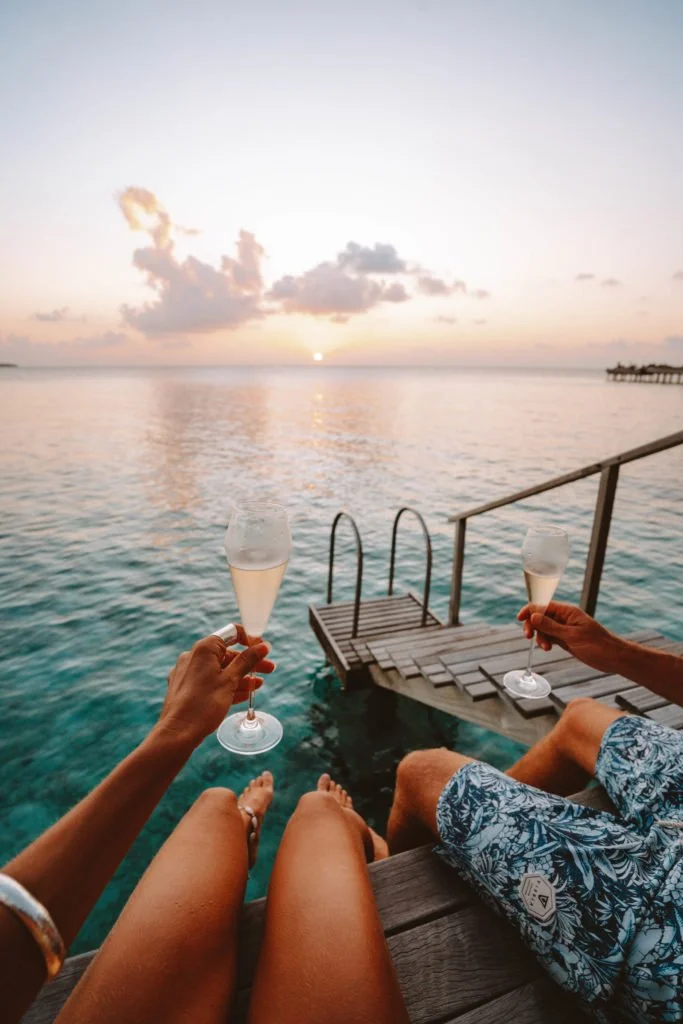 Two people sitting on a dock by the water, holding glasses of champagne, with a sunset in the background.