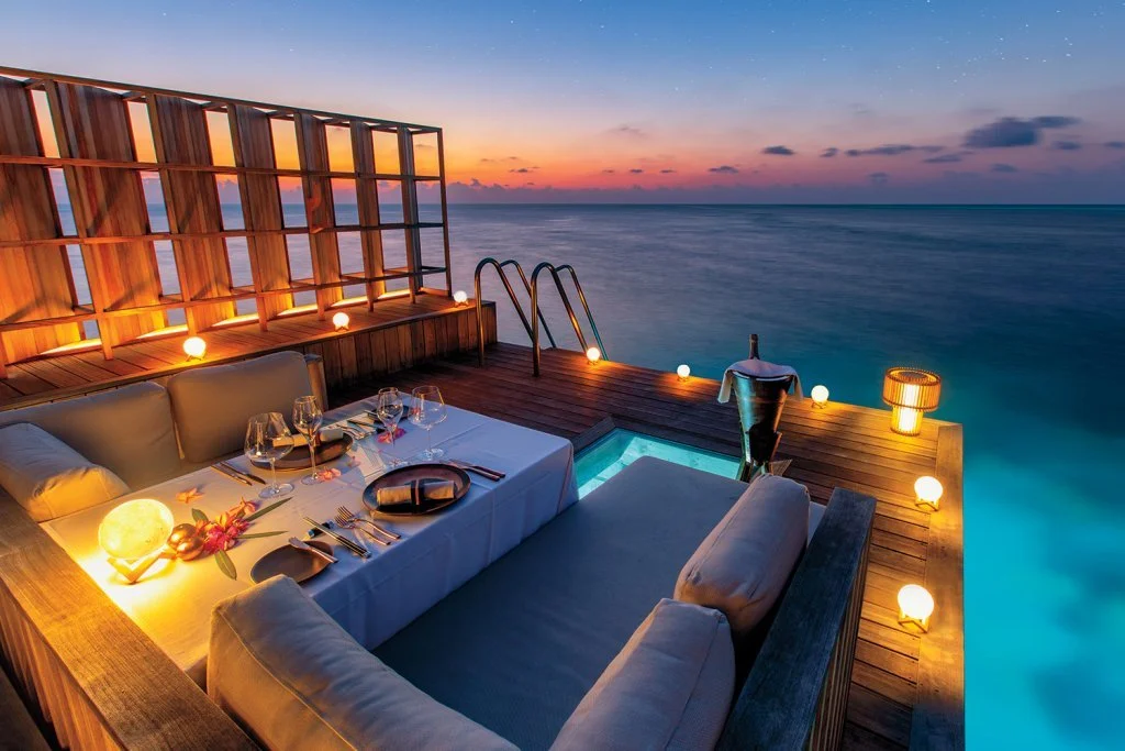 A cozy outdoor dining area on a wooden deck overlooking the ocean during sunset, illuminated by warm string lights, with a table set for dinner with wine glasses, cutlery, and decorative lighting.