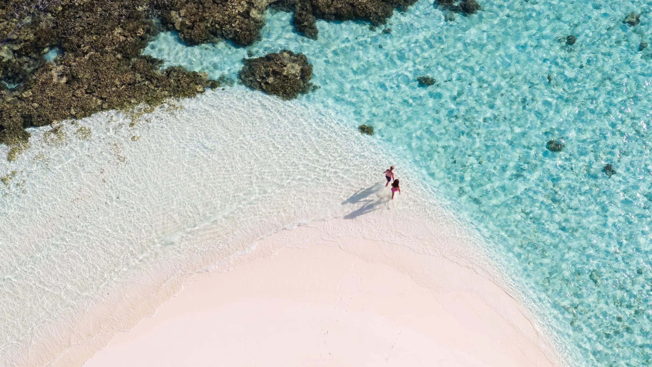 An aerial view of two children walking on a sandy beach near turquoise water with rocks.