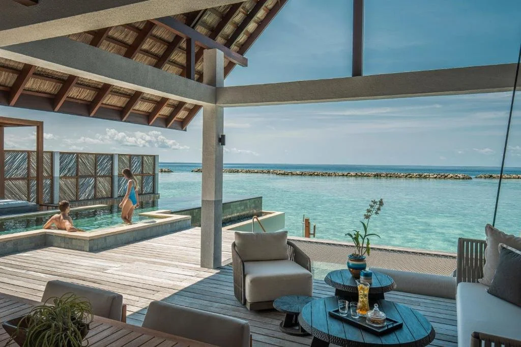 A view of a seaside resort with a pool and outdoor sitting area, where two children are playing in the pool, overlooking a clear blue ocean with a breakwater in the distance.