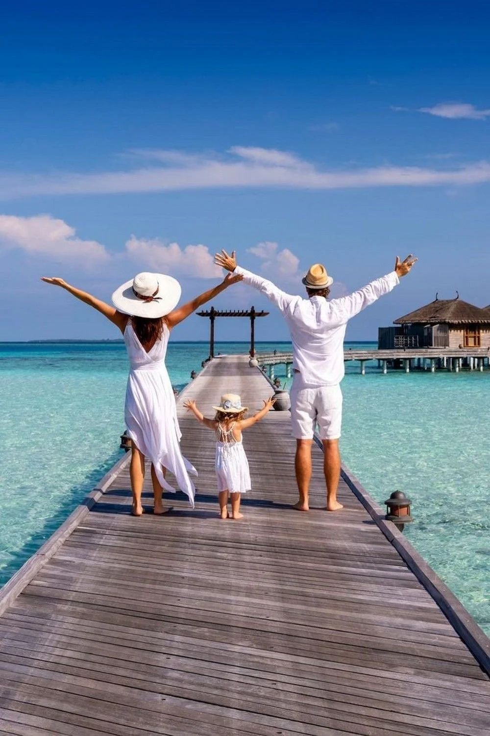 A family of three standing on a wooden pier at a tropical beach, dressed in white, with arms raised, facing the ocean, with overwater bungalows in the background.