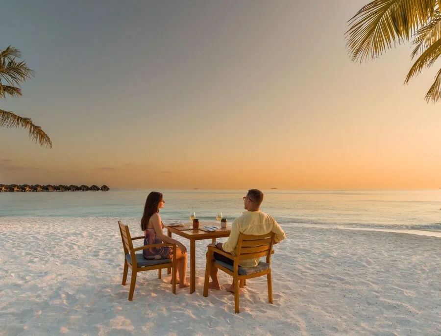 A man and woman sitting at a wooden table with drinks on a beach at sunset, surrounded by palm trees.