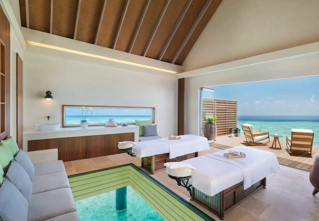Luxury spa room with two massage tables, a view of the ocean through a large window and balcony, and a built-in pool or hot tub.