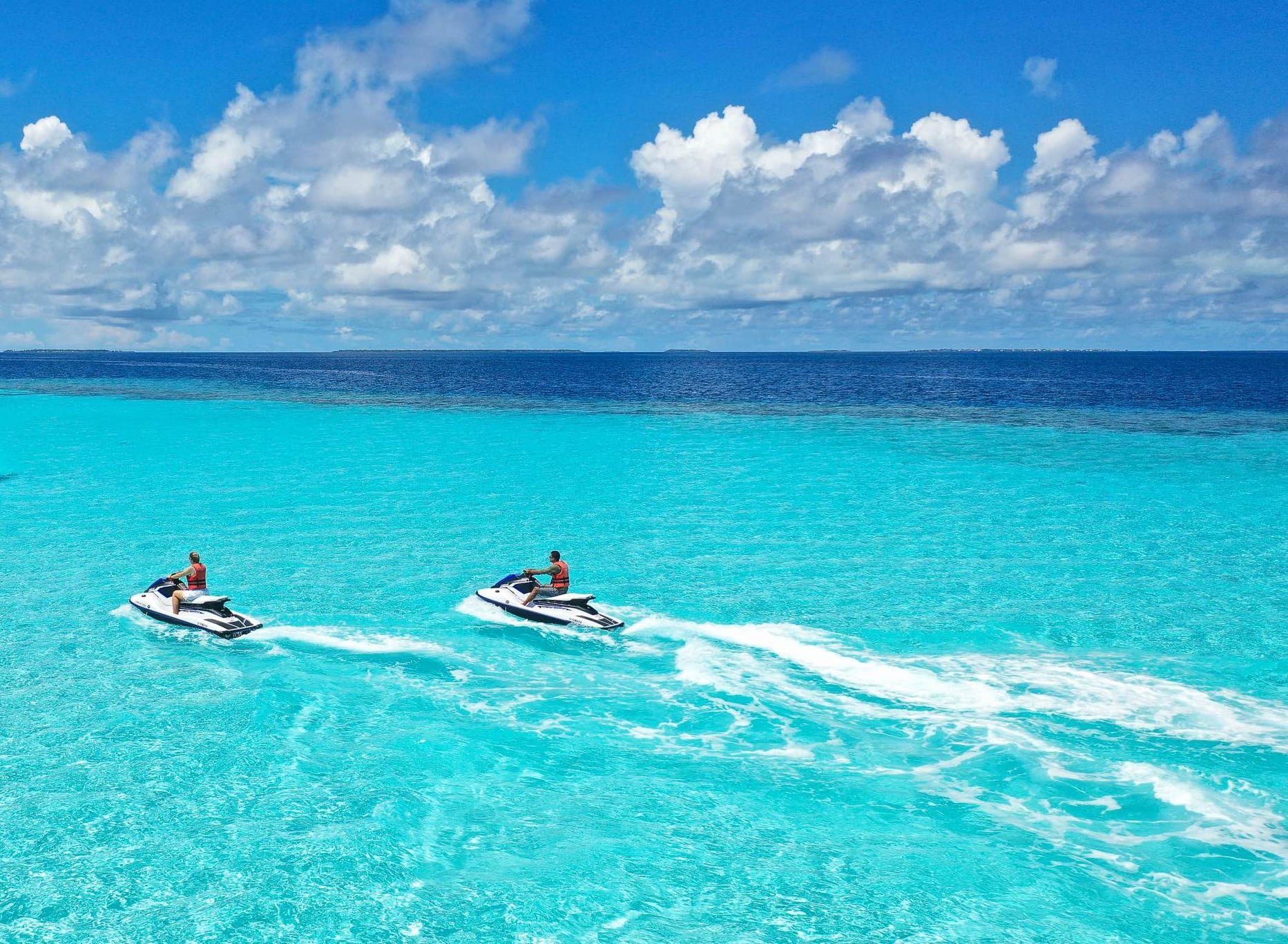 Two people riding jet skis on turquoise ocean water with a cloudy sky and distant land in the background.