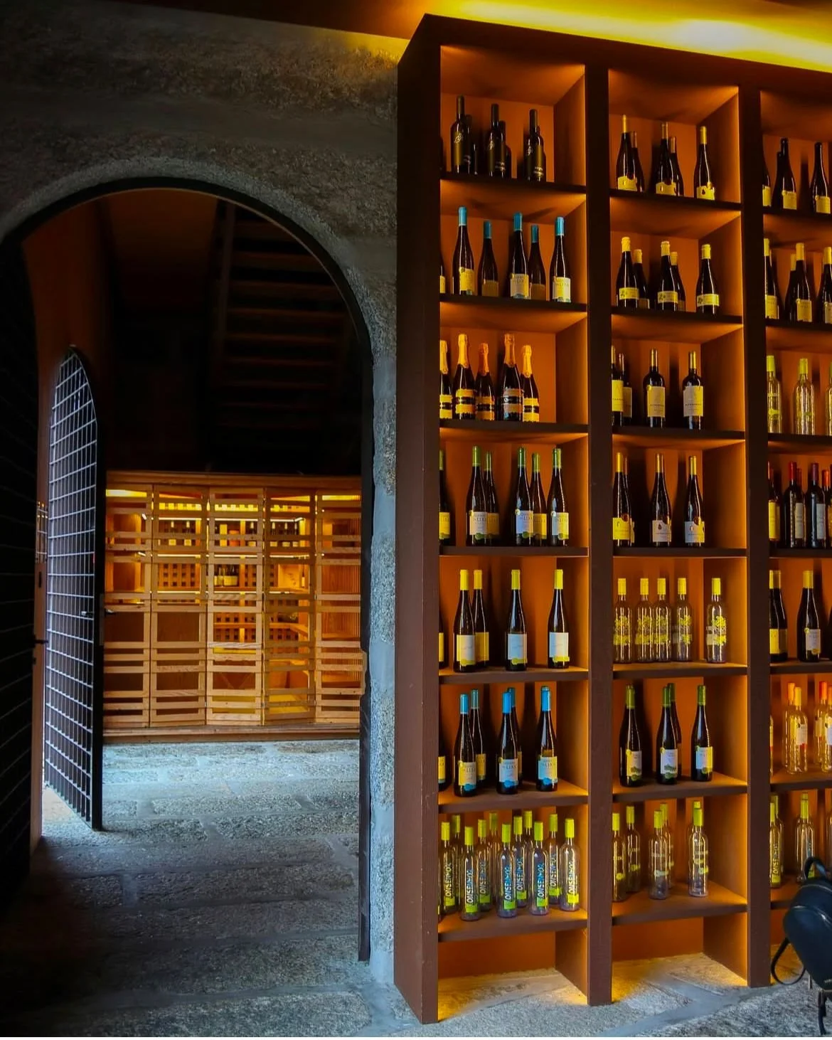 Estante de madeira com garrafas de vinho decorativas ao interior de uma adega, com iluminação quente. Ao lado, uma porta de ferro se abre para uma passagem com piso de pedra e paredes de pedra.