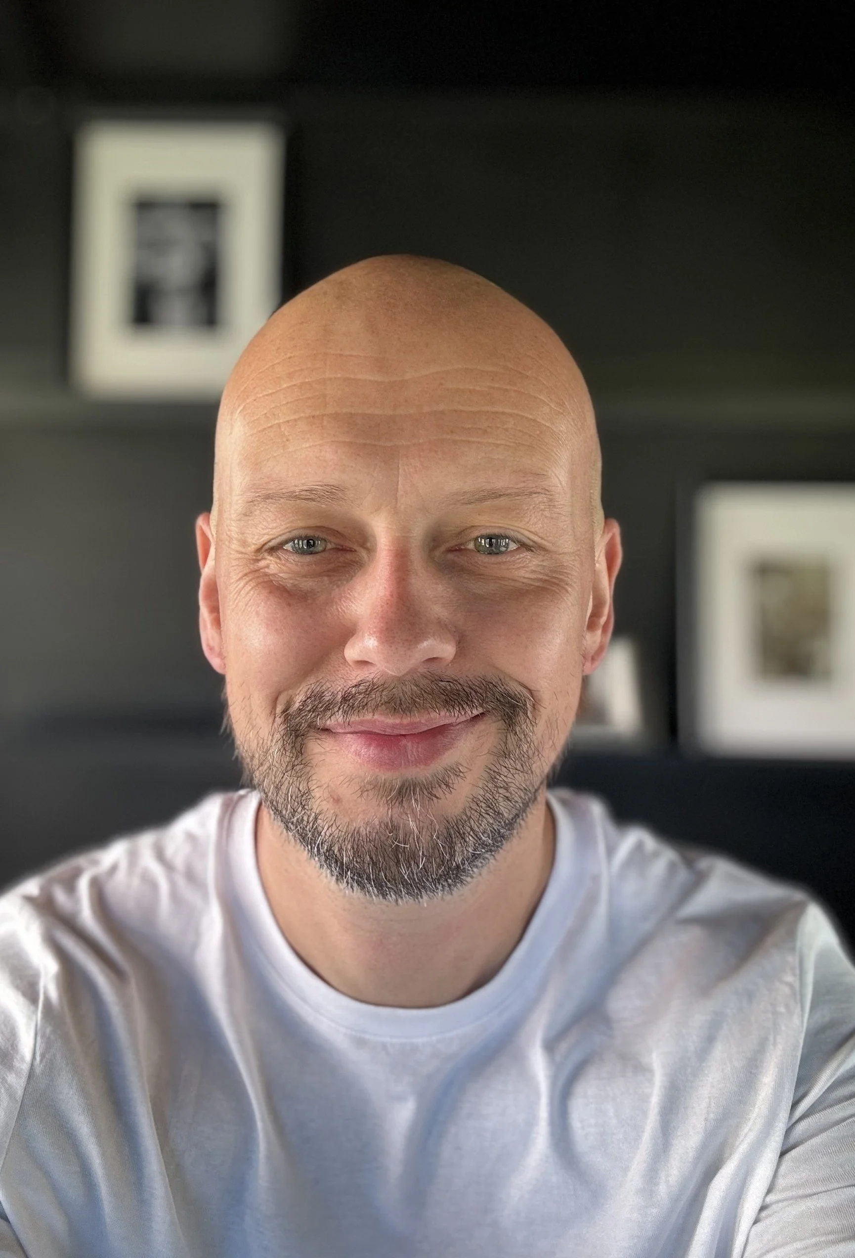 A smiling bald man with a beard and mustache wearing a white t-shirt, taking a selfie indoors with a dark background and framed pictures on the wall.