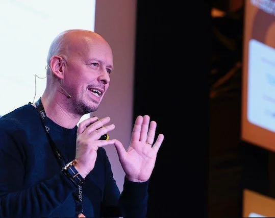 A man with a bald head and goatee speaking or presenting, gesturing with his hands, wearing a black shirt and a lanyard, in front of a screen or presentation background.
