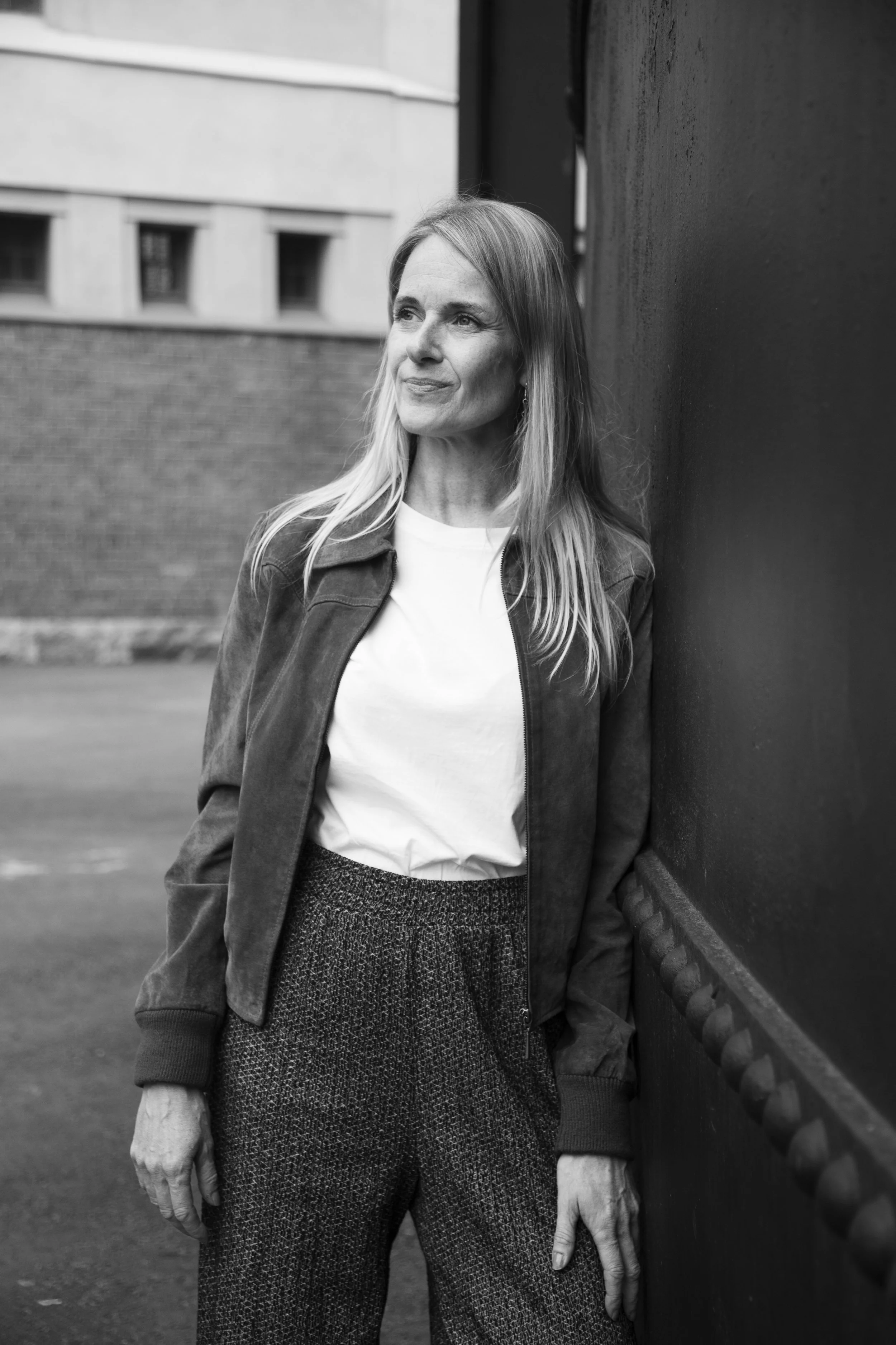 Mustavalkokuva naisesta seisomassa ulkona, nojaten tiiliseinään.