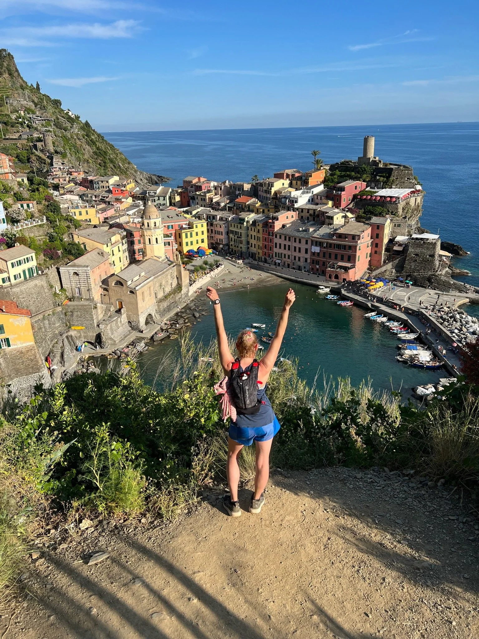 A woman with a backpack standing on a dirt trail overlooking a colorful seaside village with boats in the harbor, tall cliffs, and a castle on a rocky promontory, with the ocean in the background.