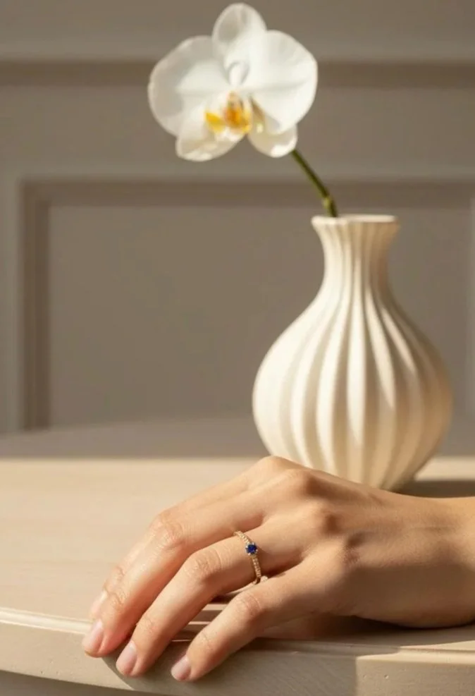 Une main portant une bague avec une pierre bleue, reposant sur une surface en bois, avec une vase blanche à rayures ondulées contenant une orchidée blanche en arrière-plan.