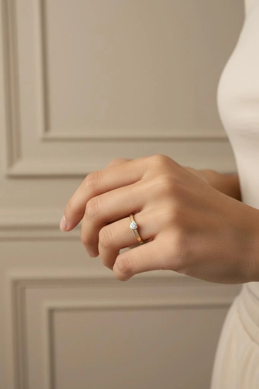 Une main portant une bague en or avec une pierre précieuse, probablement un diamant, sur le doigt annulaire, contre un fond beige avec des moulures murales.