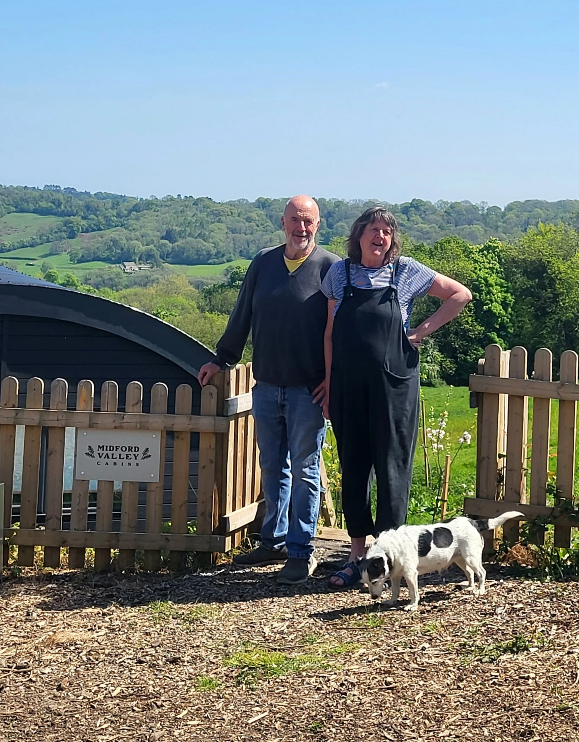 Two people standing outdoors behind a wooden fence with a pasture and green rolling hills in the background, a black and white dog is on the ground, and a sign on the fence that reads "Midford Valley Cabins."