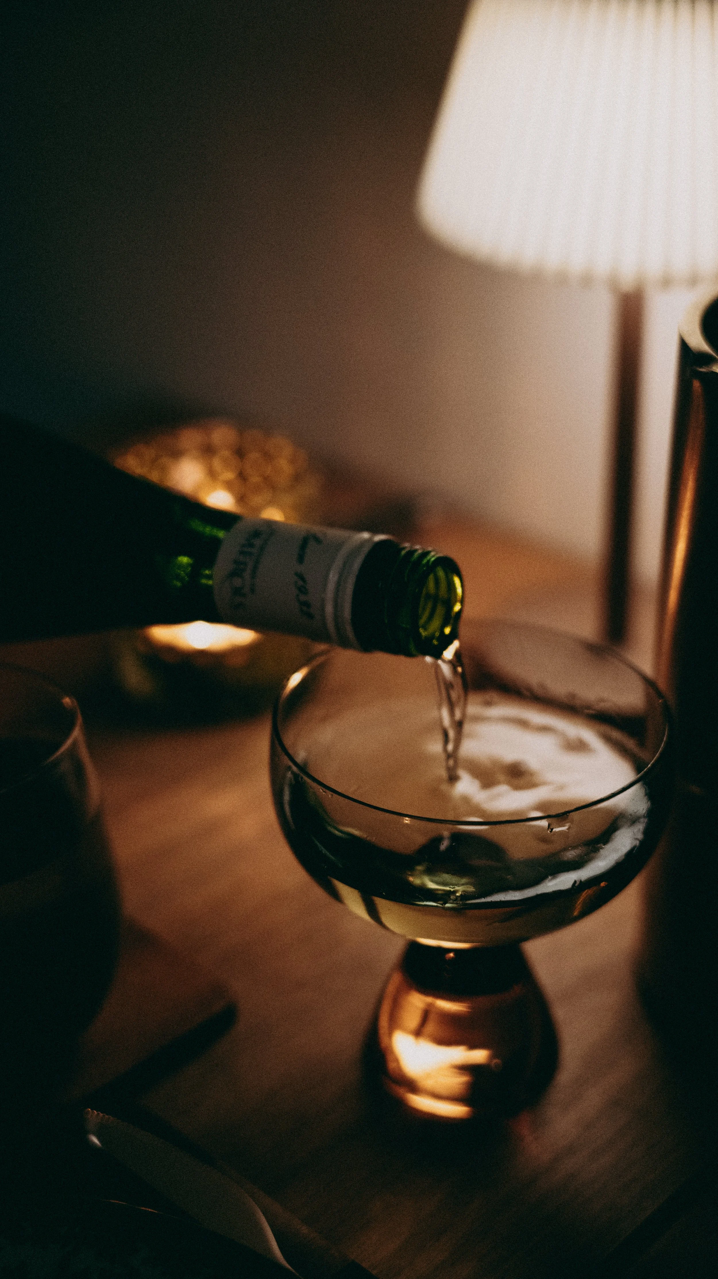 A bottle of white wine with a white label is pouring wine into a glass on a wooden table. There is a lamp and a decorative dish in the background.