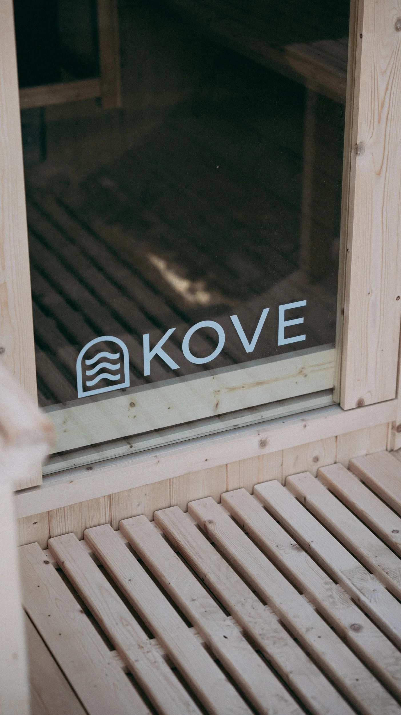 Inside a sauna with light wood benches and wall, and a glass door with the logo 'KOVE' and a water wave icon.