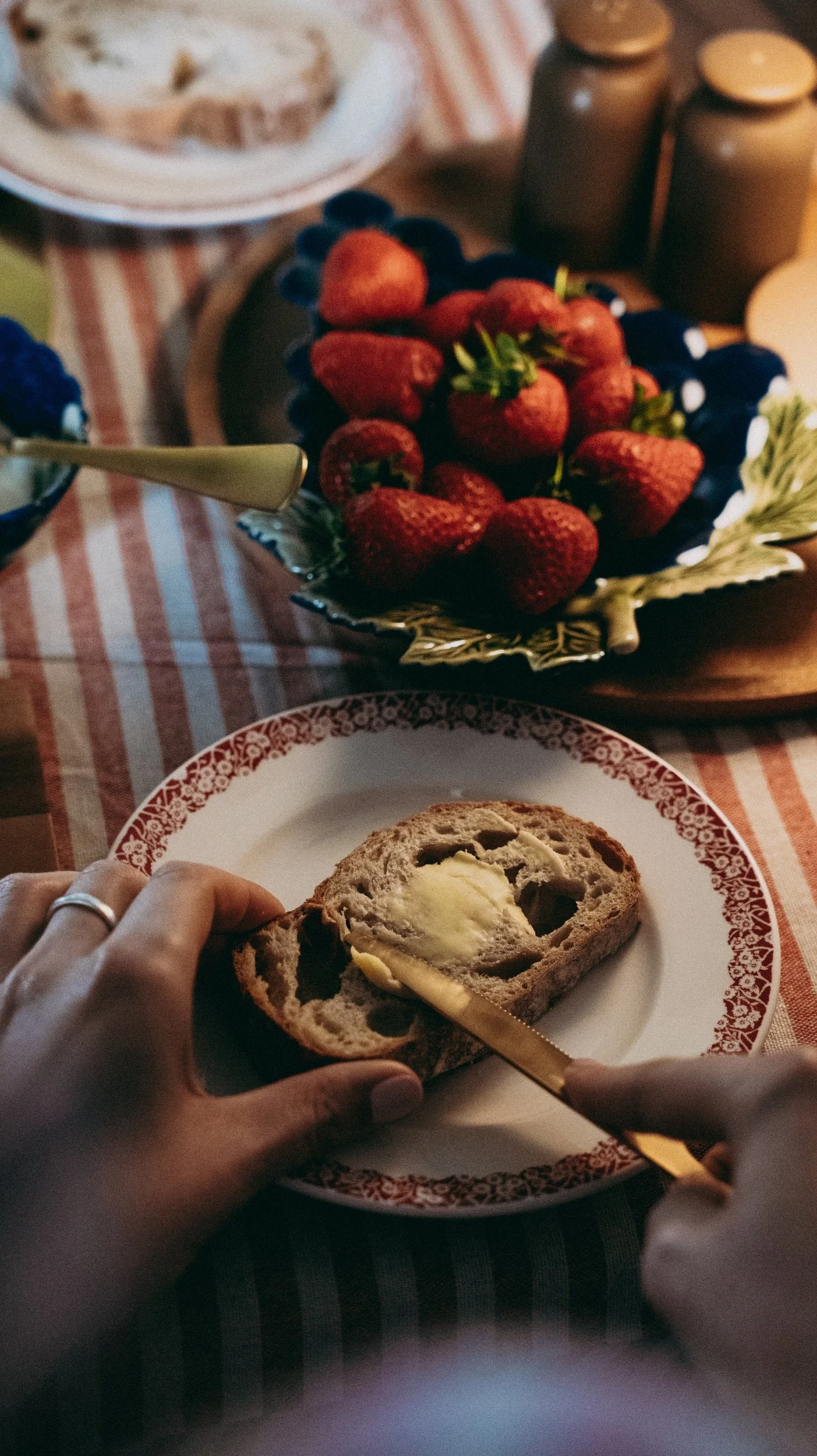 Person spreading butter on a slice of bread with a gold knife, with a plate of strawberries and a bowl of blueberries in the background.