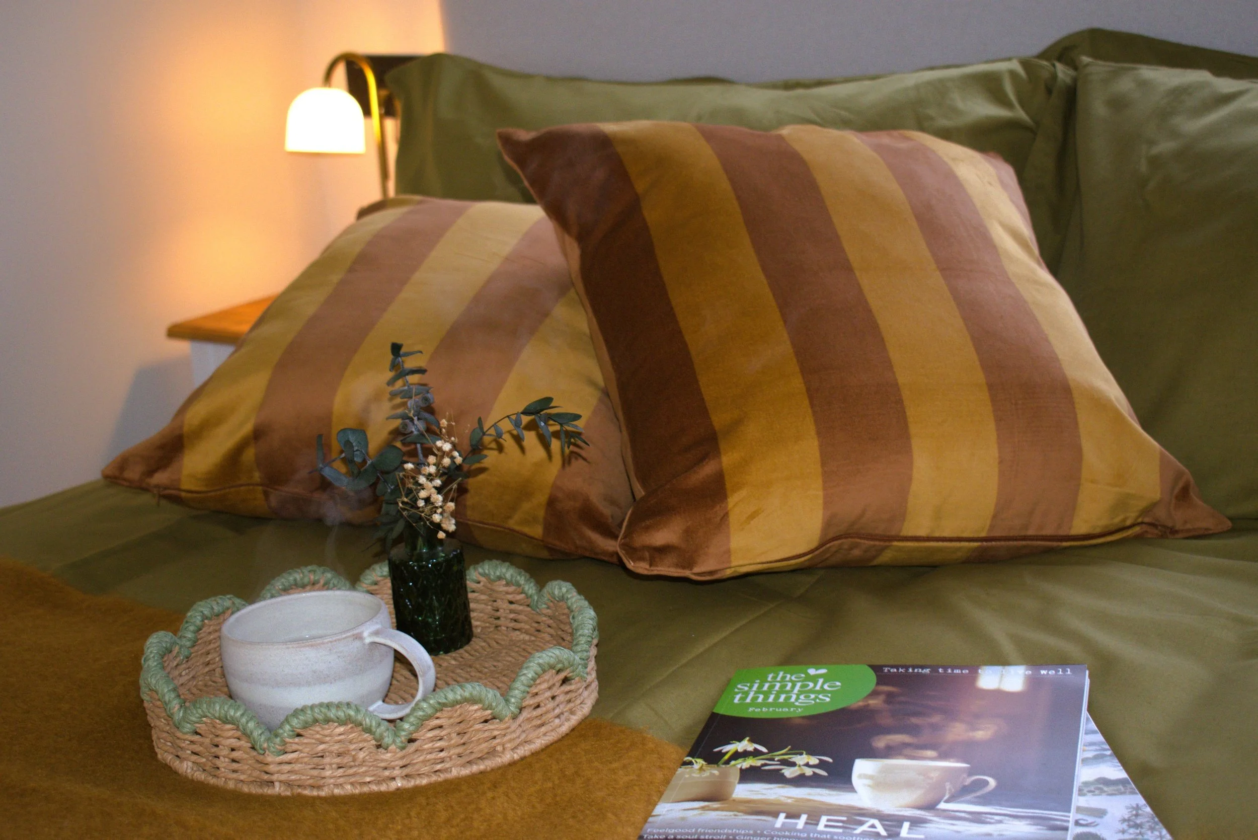 A cozy bedroom corner with green and brown striped pillows on a bed, a small black vase with eucalyptus leaves, a wicker tray, a white mug, and a magazine titled "the simple things" on a wooden surface, illuminated by a wall-mounted lamp.