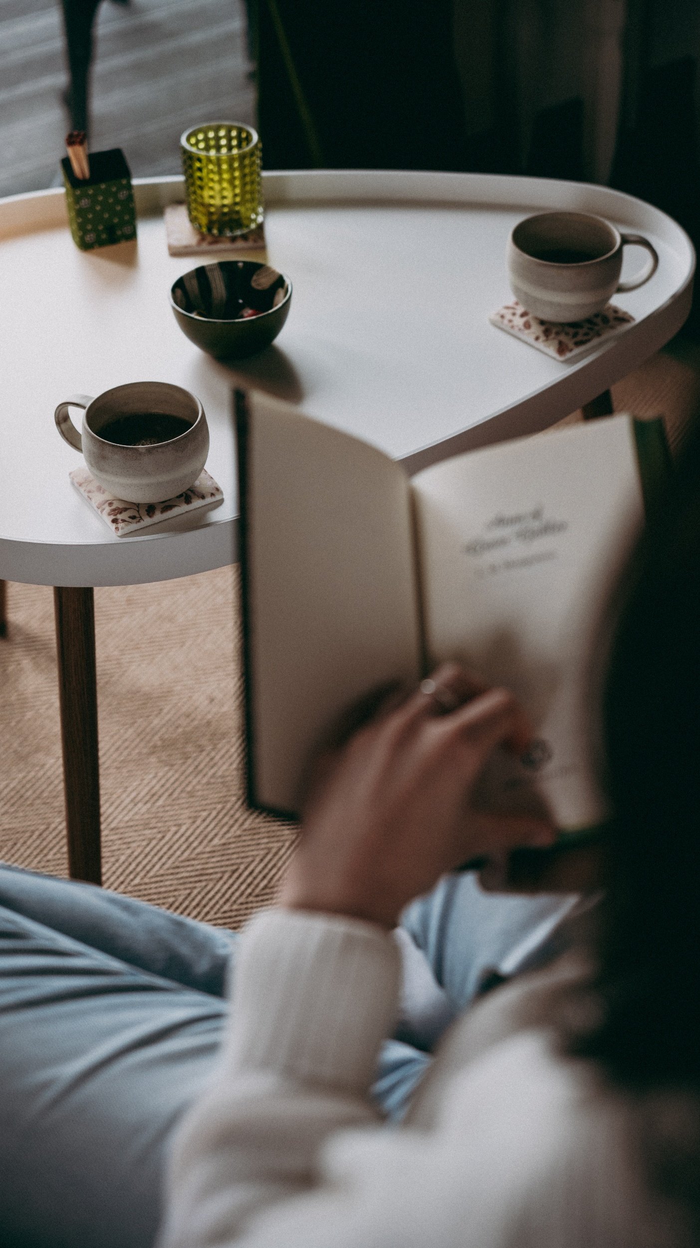 A person reading a book while sitting. In the background, there's a white table with two coffee cups, a green bowl.