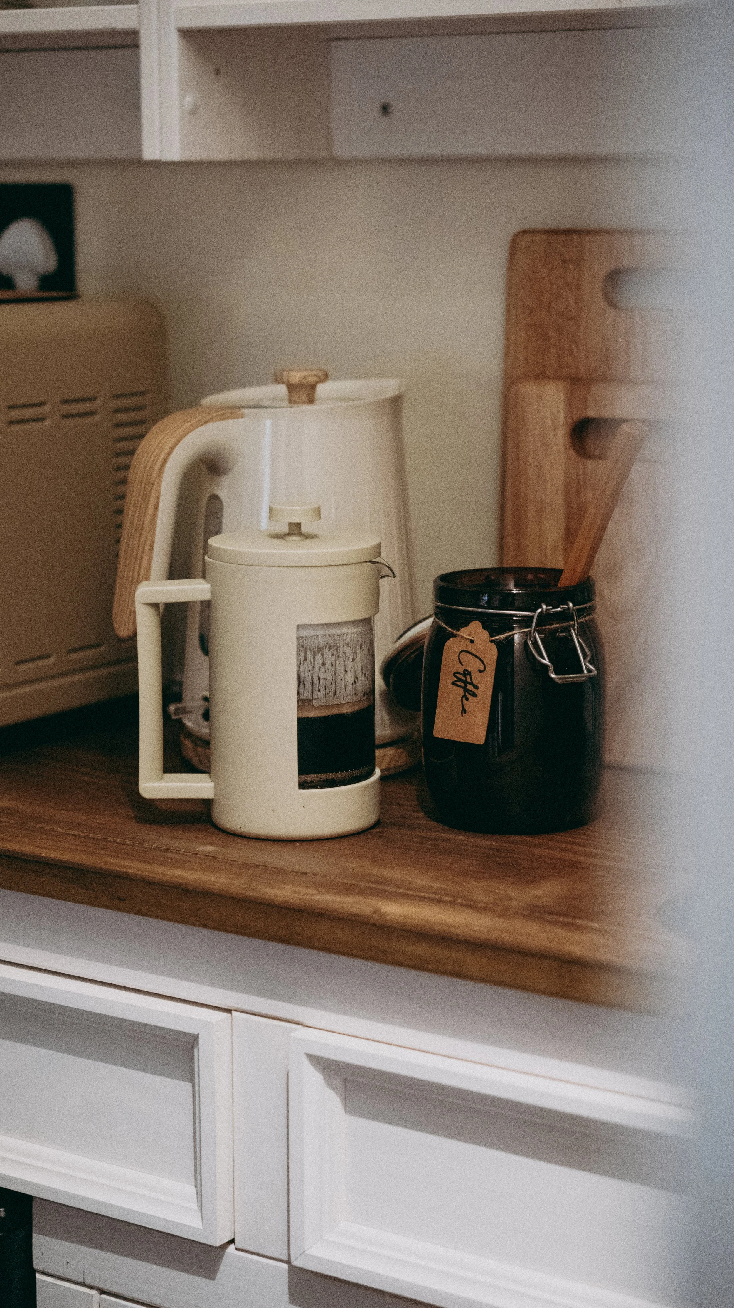 Kitchen countertop with a white French press, a black jar with a tag that says 'Coffee', and a wooden cutting board behind them.