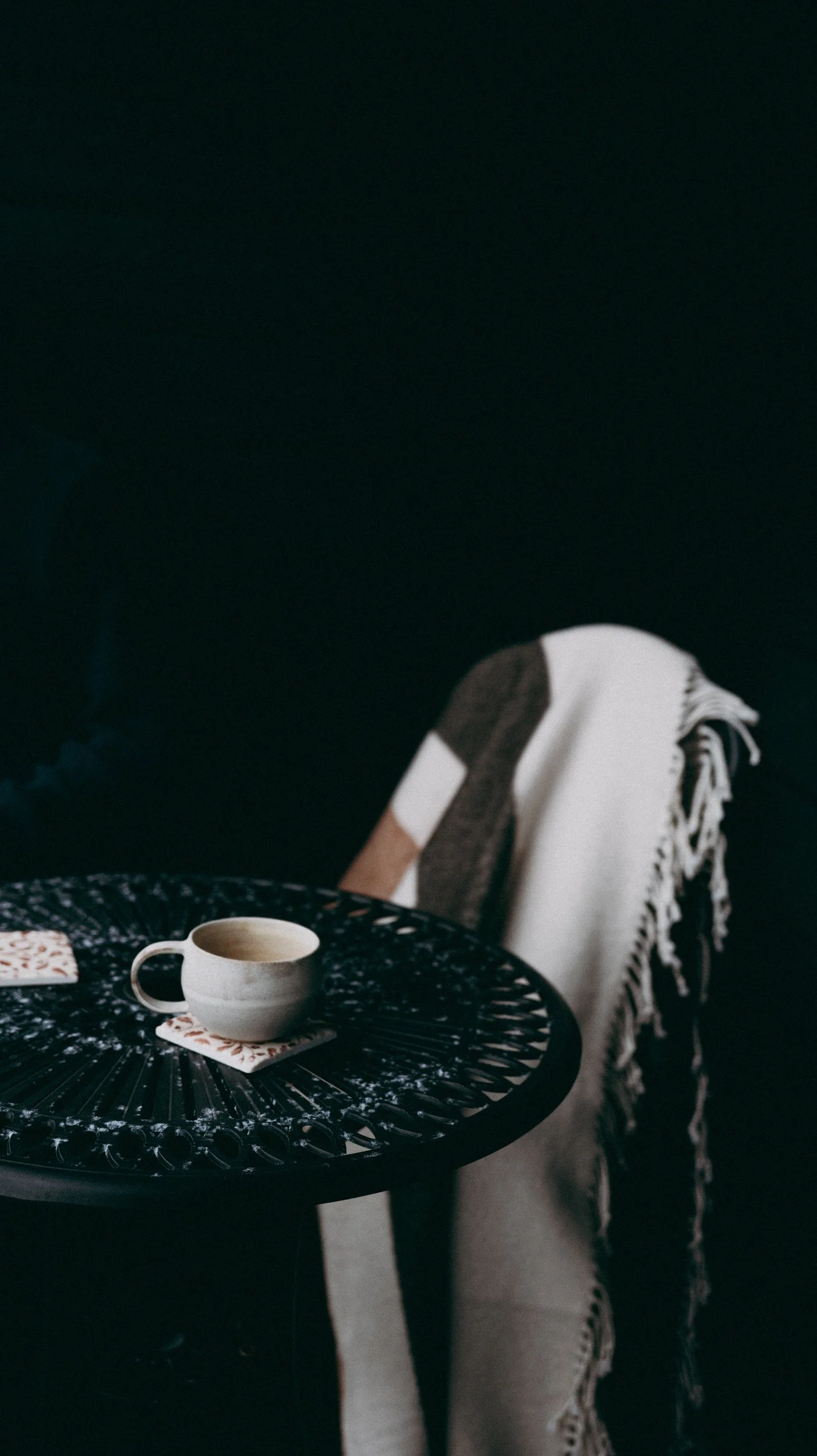 A black outdoor table with a coffee cup and a small plate. A beige and gray blanket is draped over a black chair next to the table, against a dark background.