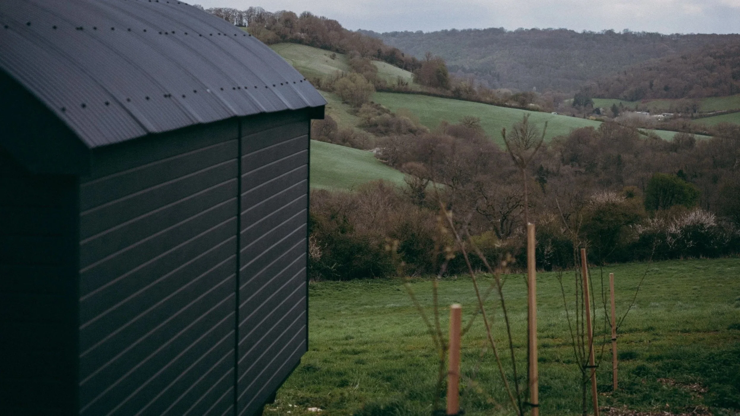 A black cabin with a curved roof situated on a lush green hill, with rolling hills and trees in the background under an overcast sky.