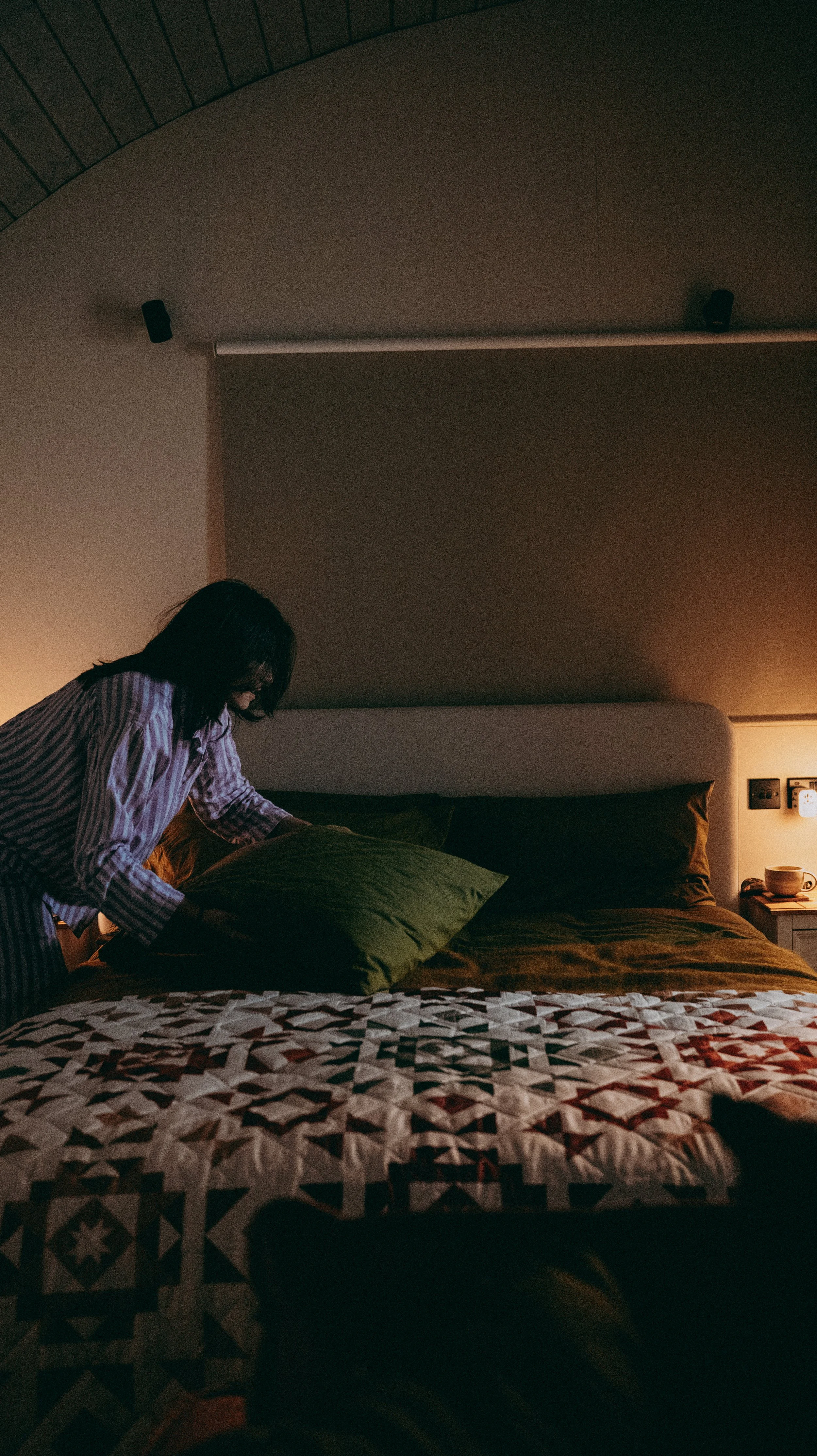 A person with dark hair, wearing striped pajamas, making the bed in a dimly lit bedroom with green bedding and green pillows. There is a bedside table with a mug and a small lamp on it.