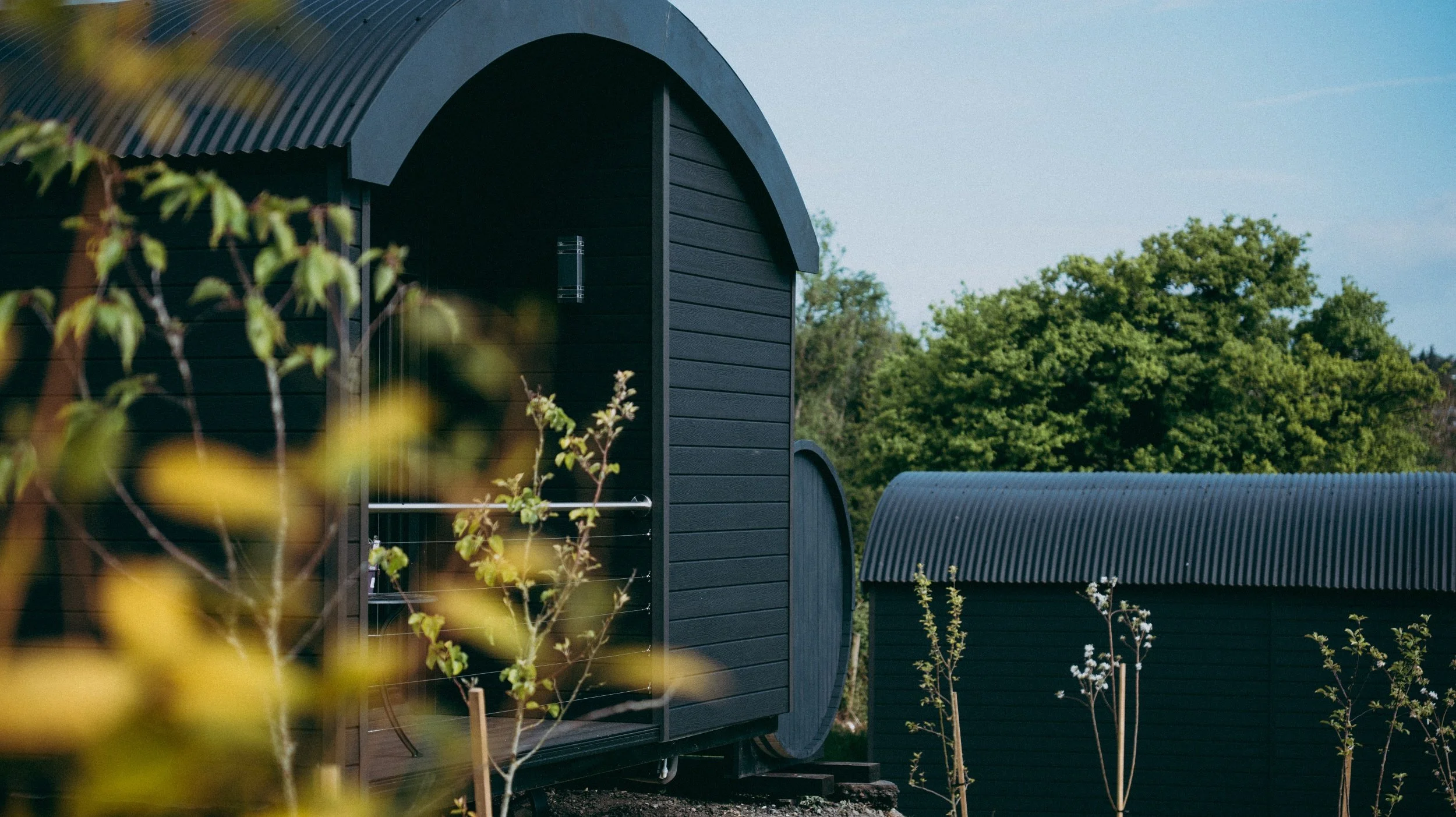 Black modern cabins with curved roof, surrounded by green trees and plants, under a clear blue sky.