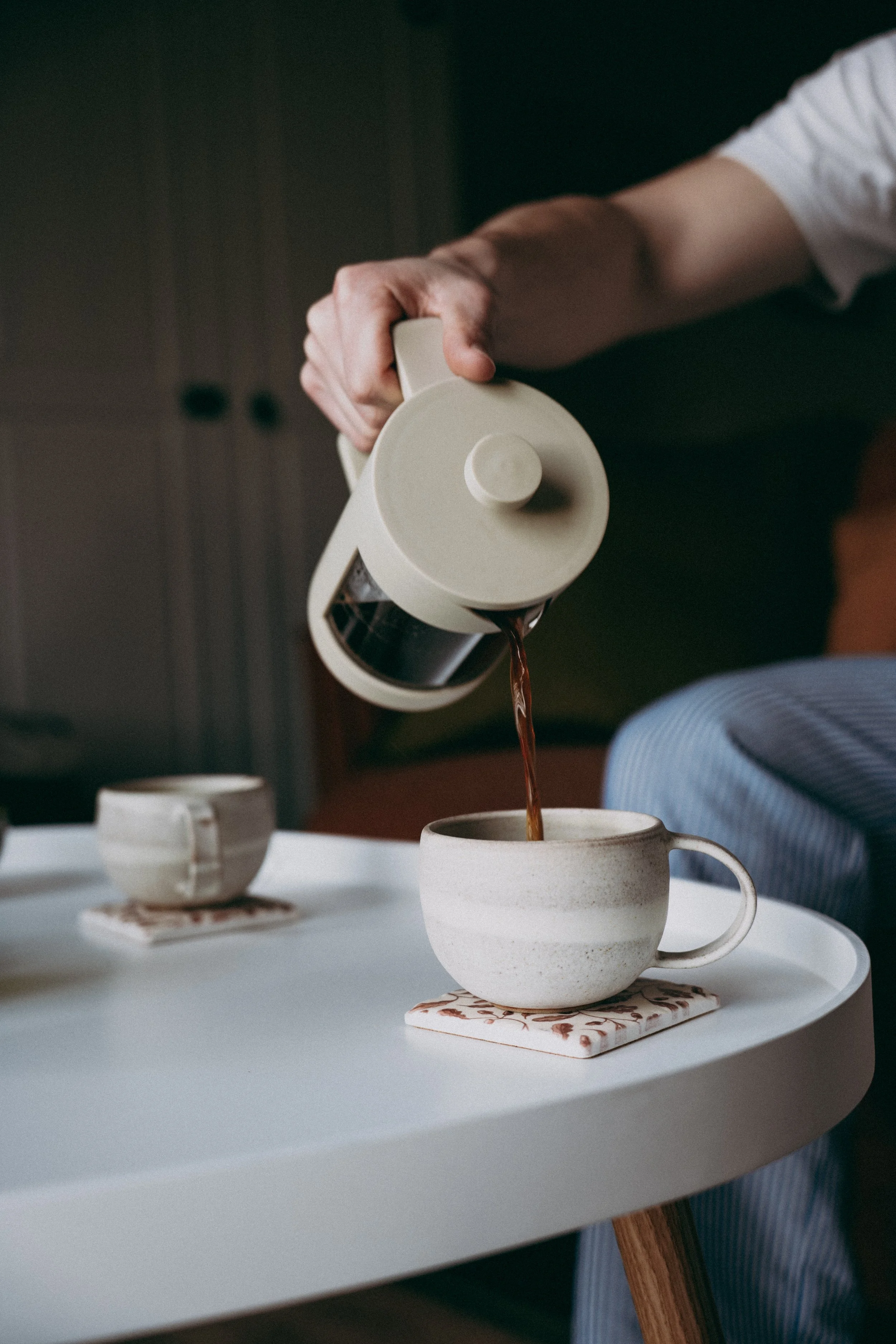 Person pouring coffee from a cream-colored coffee pot into a mug on a white table, with two other mugs and a coaster nearby, in a cozy setting.