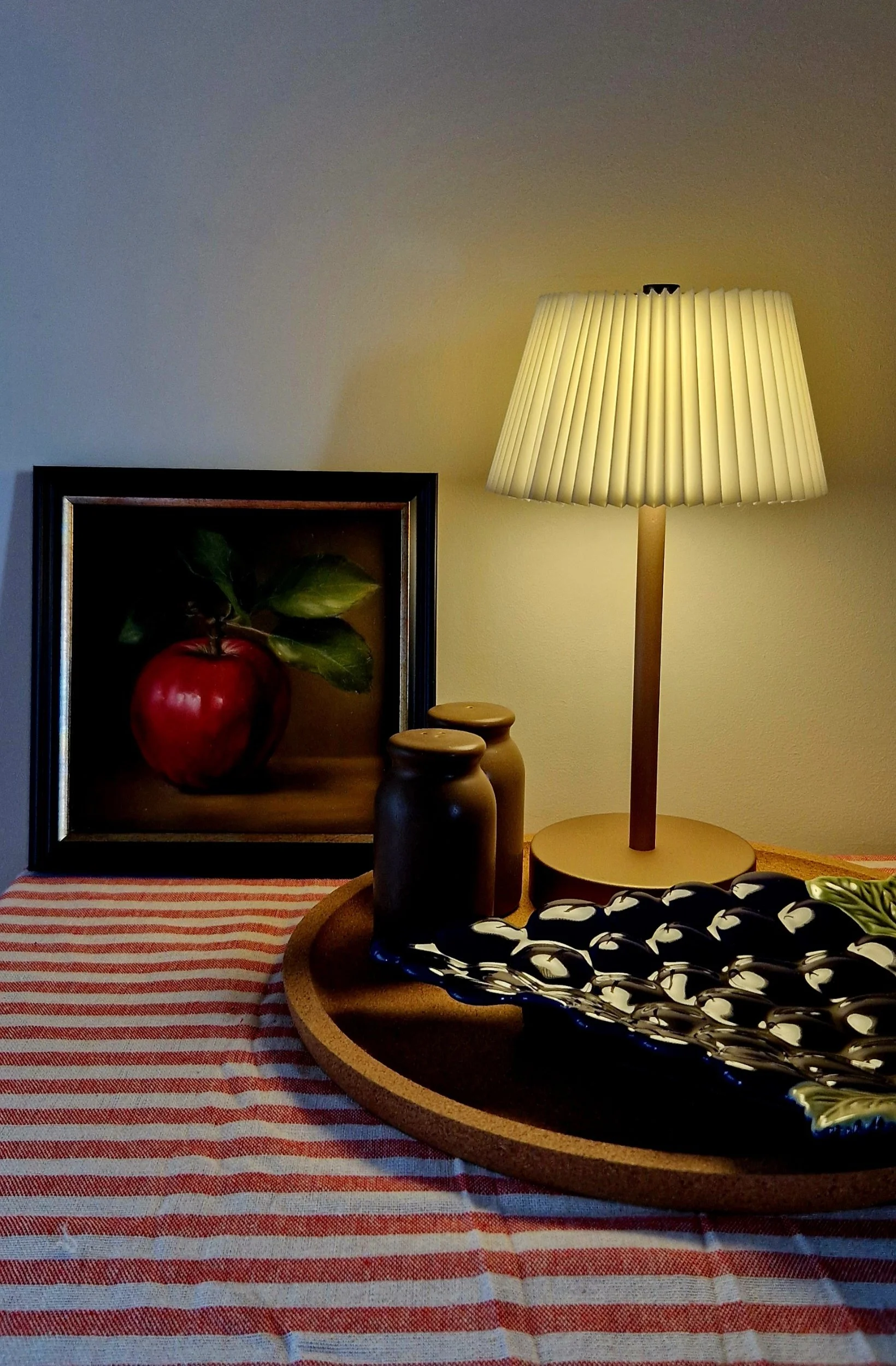 A cozy table setting with a striped tablecloth, a decorative bowl with a leaf pattern, a table lamp with a pleated shade, framed apple and leaf artwork.