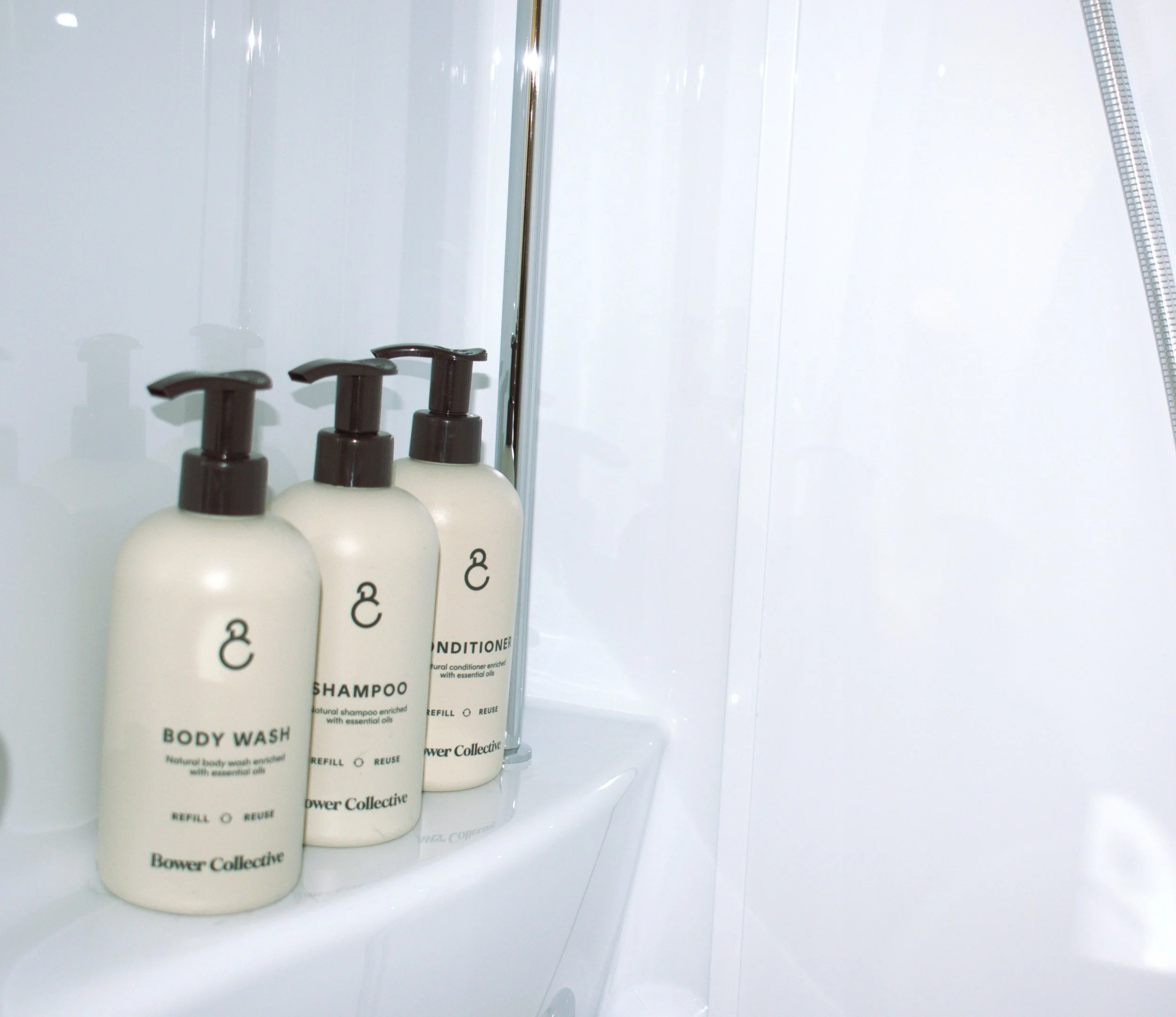 Three bottles of personal care Bower Collective products on a shower shelf, labeled as shampoo, conditioner and body wash, with a shower hose visible on the right.