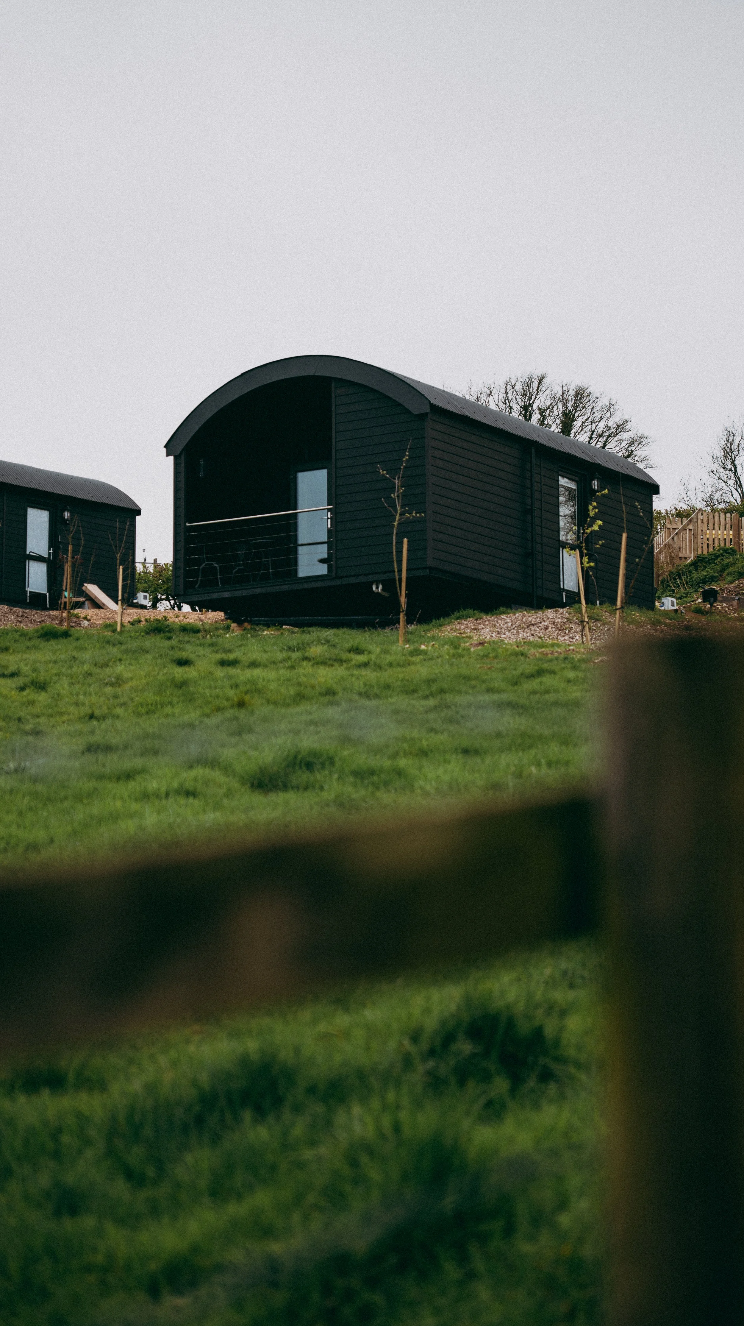 A black cabin with a curved roof and a balcony, situated on green grass with young trees around, in a rural setting.