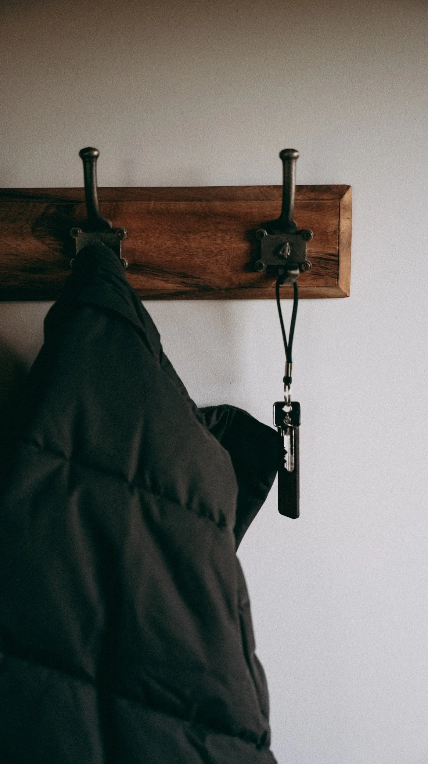 A black jacket hanging next to a set of keys with a black keychain on a wall-mounted wooden coat rack with two black hooks, against a plain white wall.