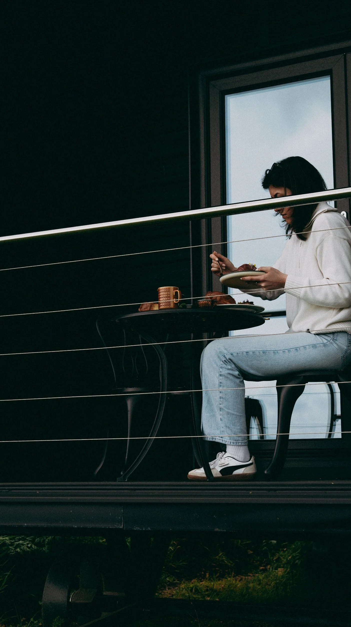A woman sitting at a table on a balcony, eating from a plate, with a window in the background. The balcony has horizontal metal railings.