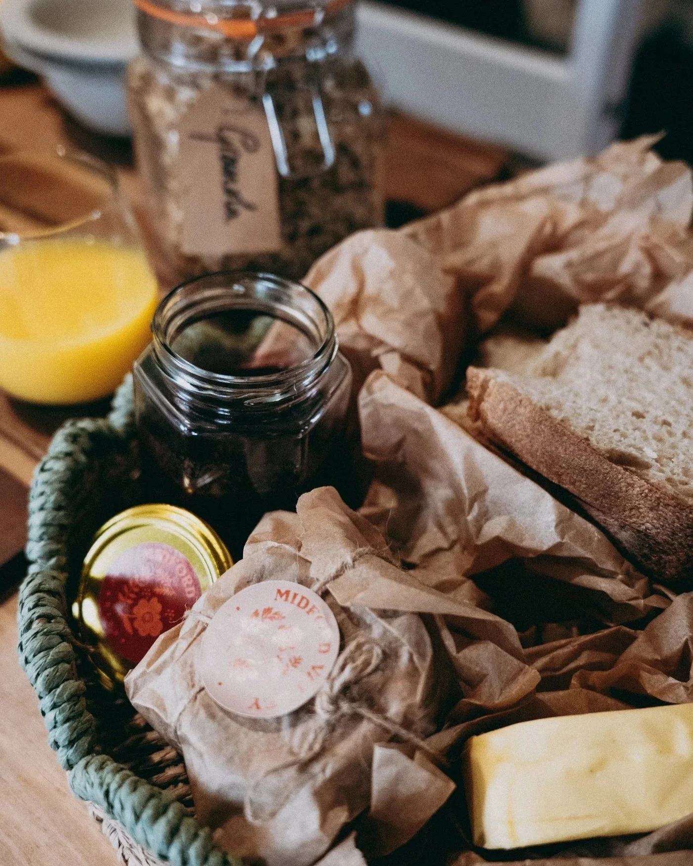 A basket containing bread, a jar of jam, a glass of orange juice, a block of butter, and other breakfast items, with some crumpled brown paper and a small jar of preserves.