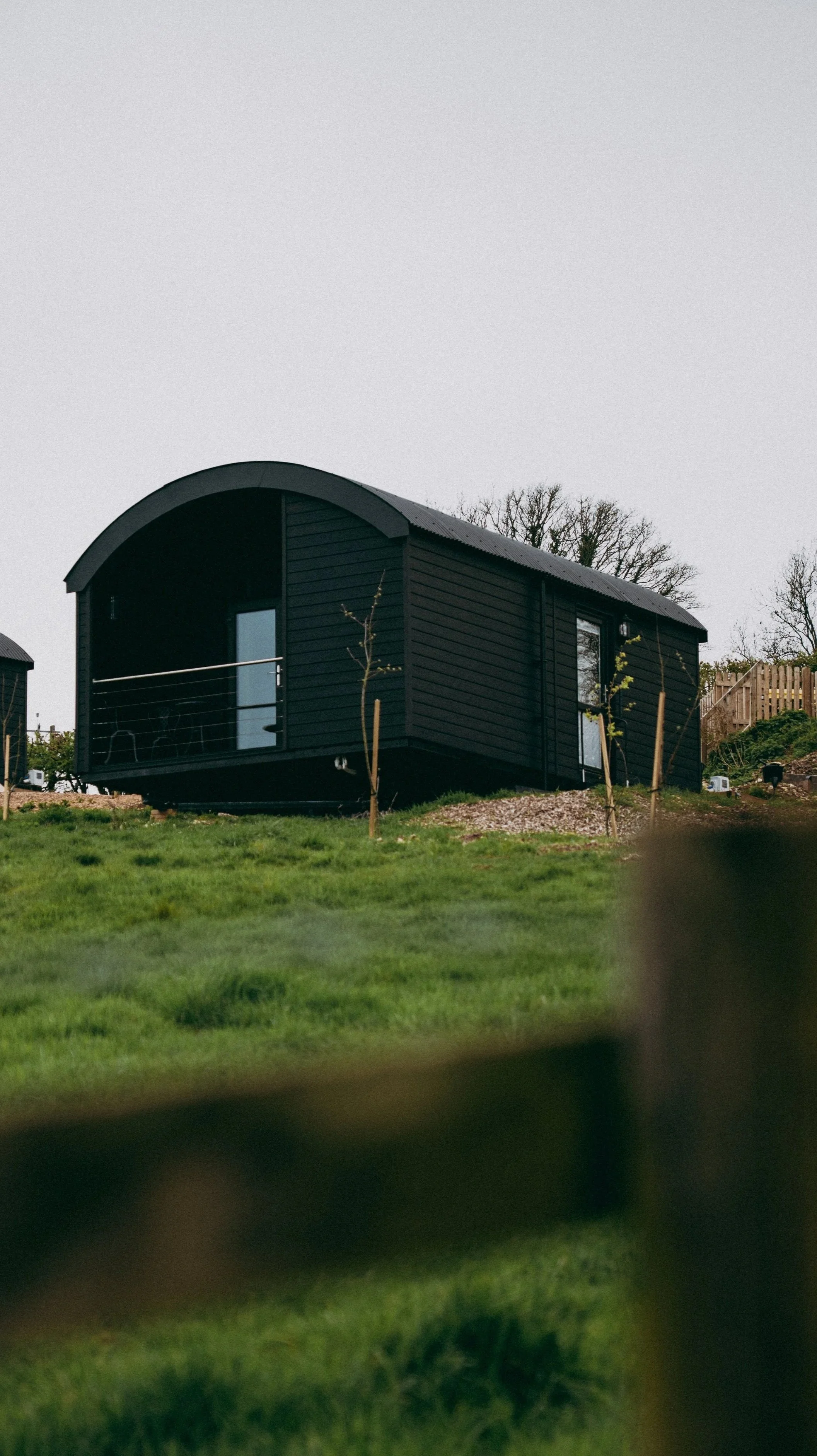 A black cabin on a grassy hill, with a small balcony, trees, and a wooden fence in the background.