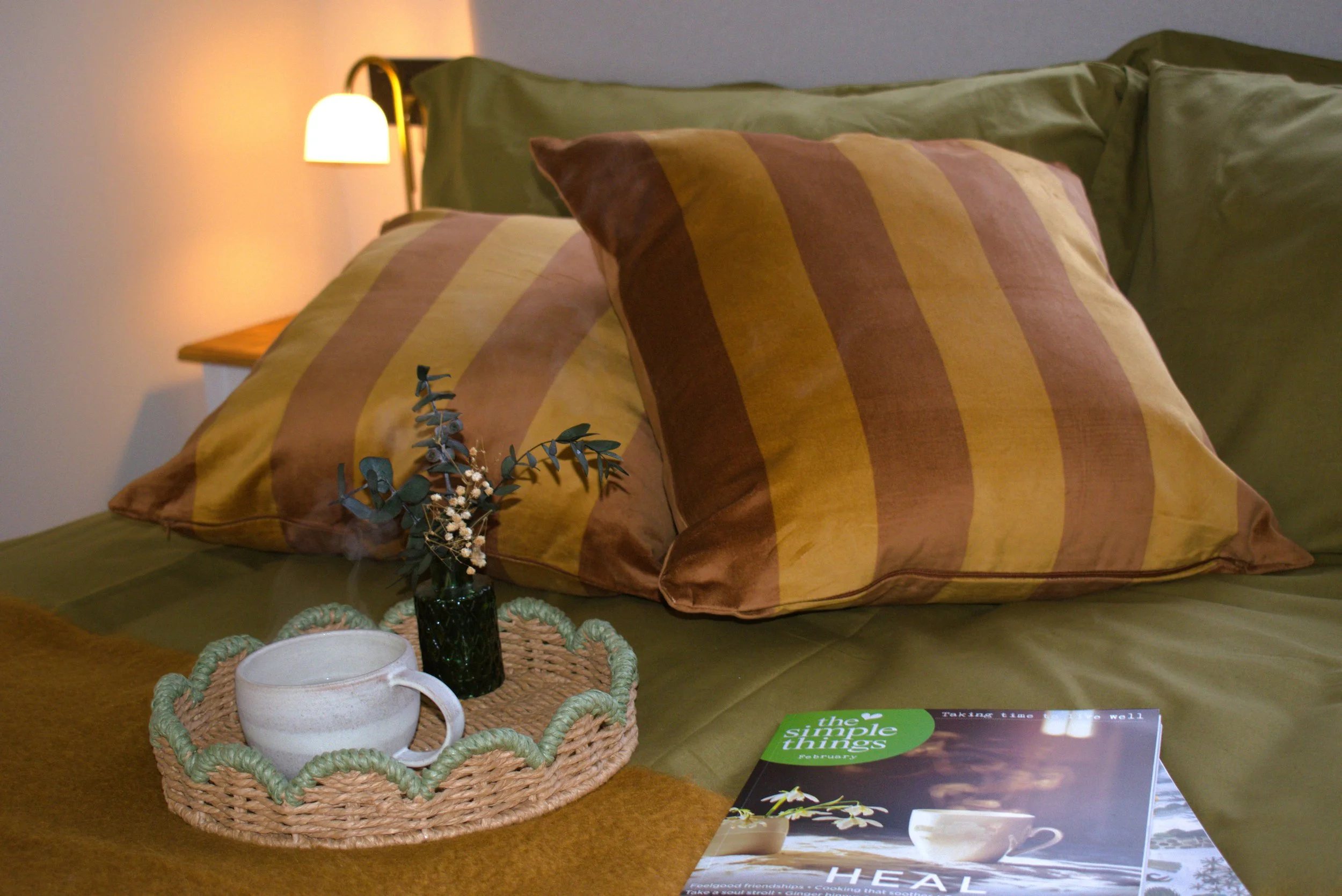 Close-up of a very comfortable bed with olive green bedding, striped brown and yellow pillows, a small bouquet of flowers in a dark vase, a white mug on a woven tray, and a magazine titled 'The Simple Things'.
