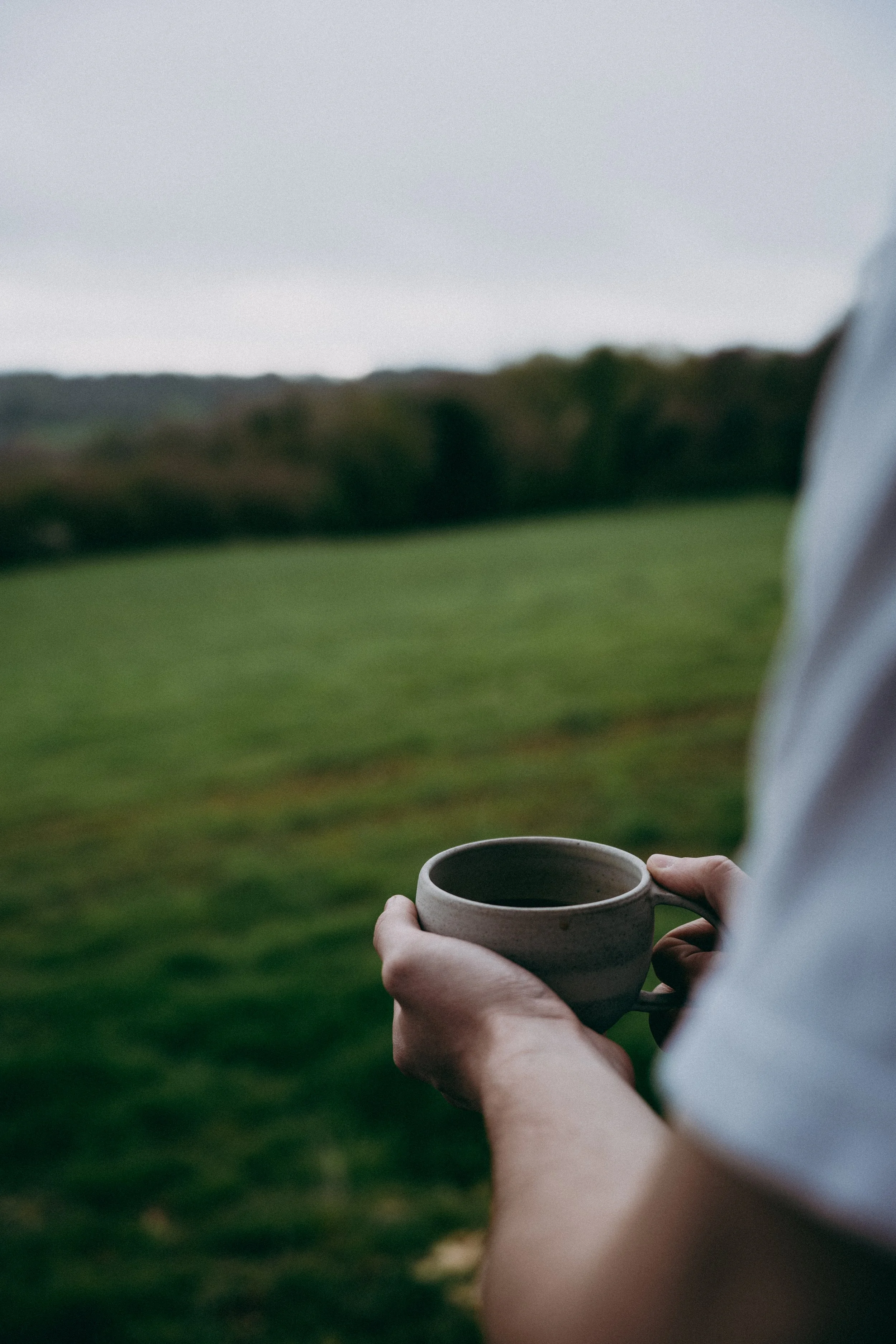 Person in a white shirt holding a beige ceramic mug filled with a hot beverage outdoors on a cloudy day, with a blurred landscape of grass and trees in the background.