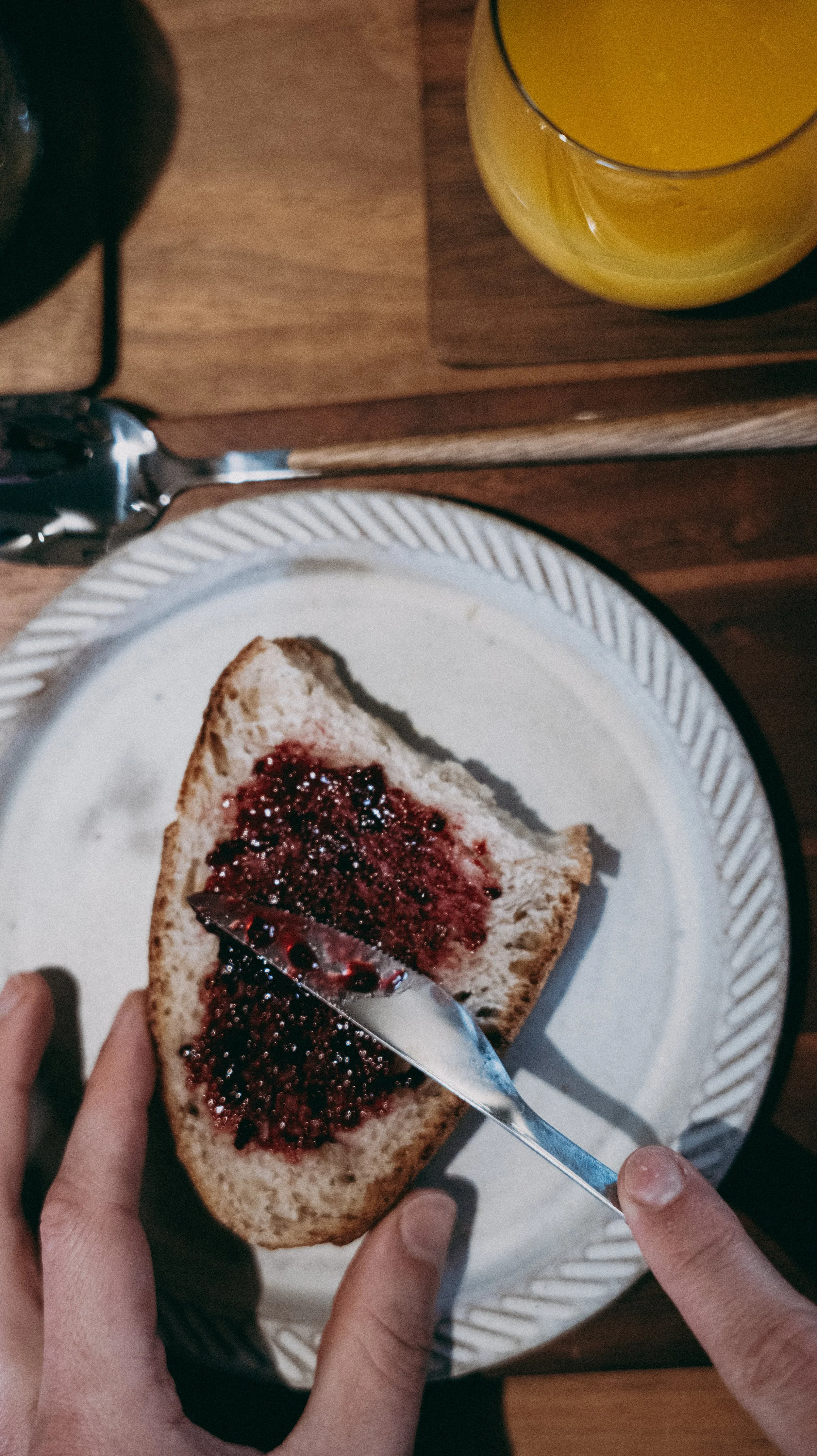 Person spreading berry jam on a slice of bread at a wooden table, with a glass of orange juice nearby.