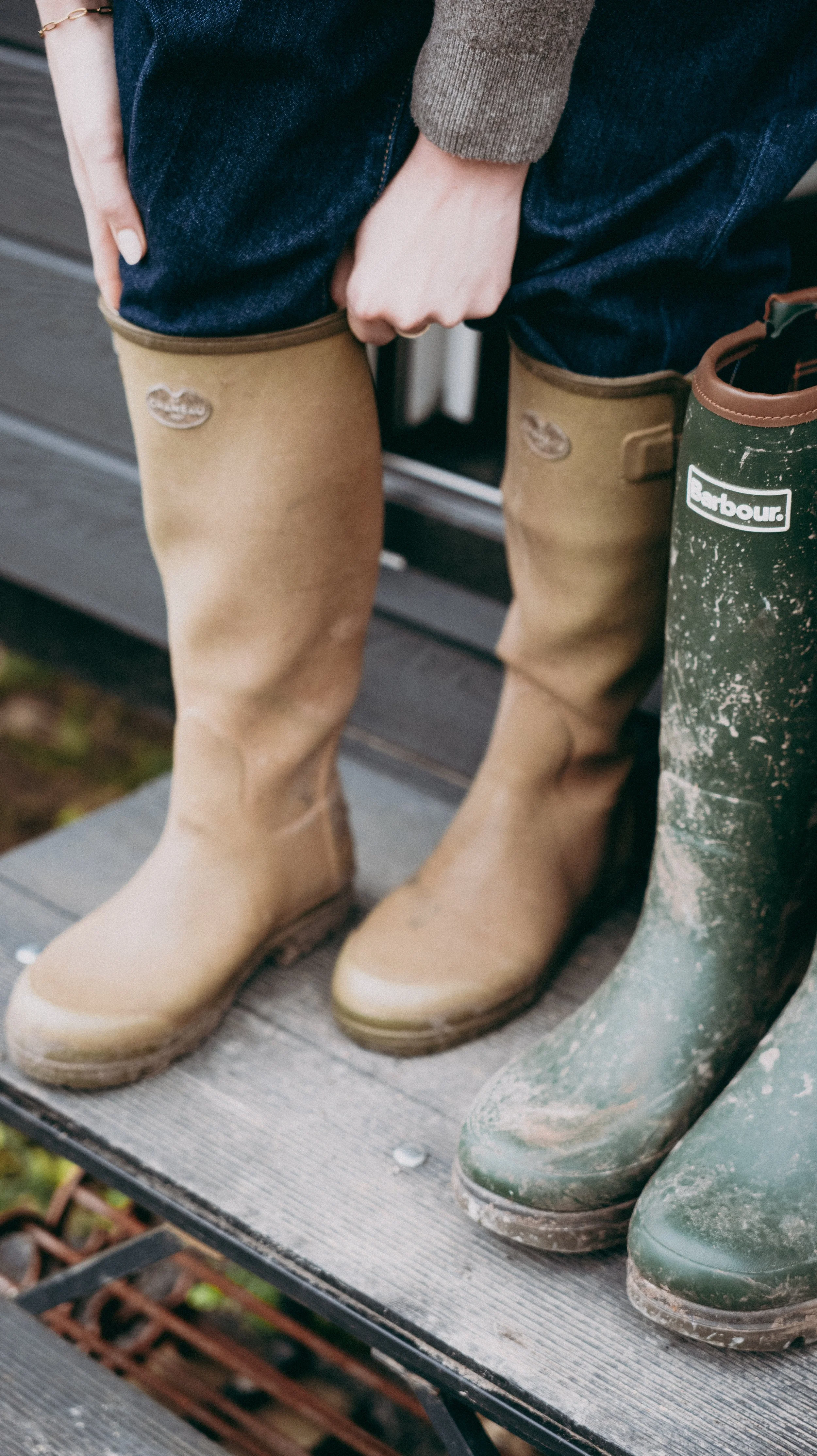 Person wearing beige Wellington boots standing on a wooden surface next to another pair of green wellington boots and a pair of dark blue jeans.