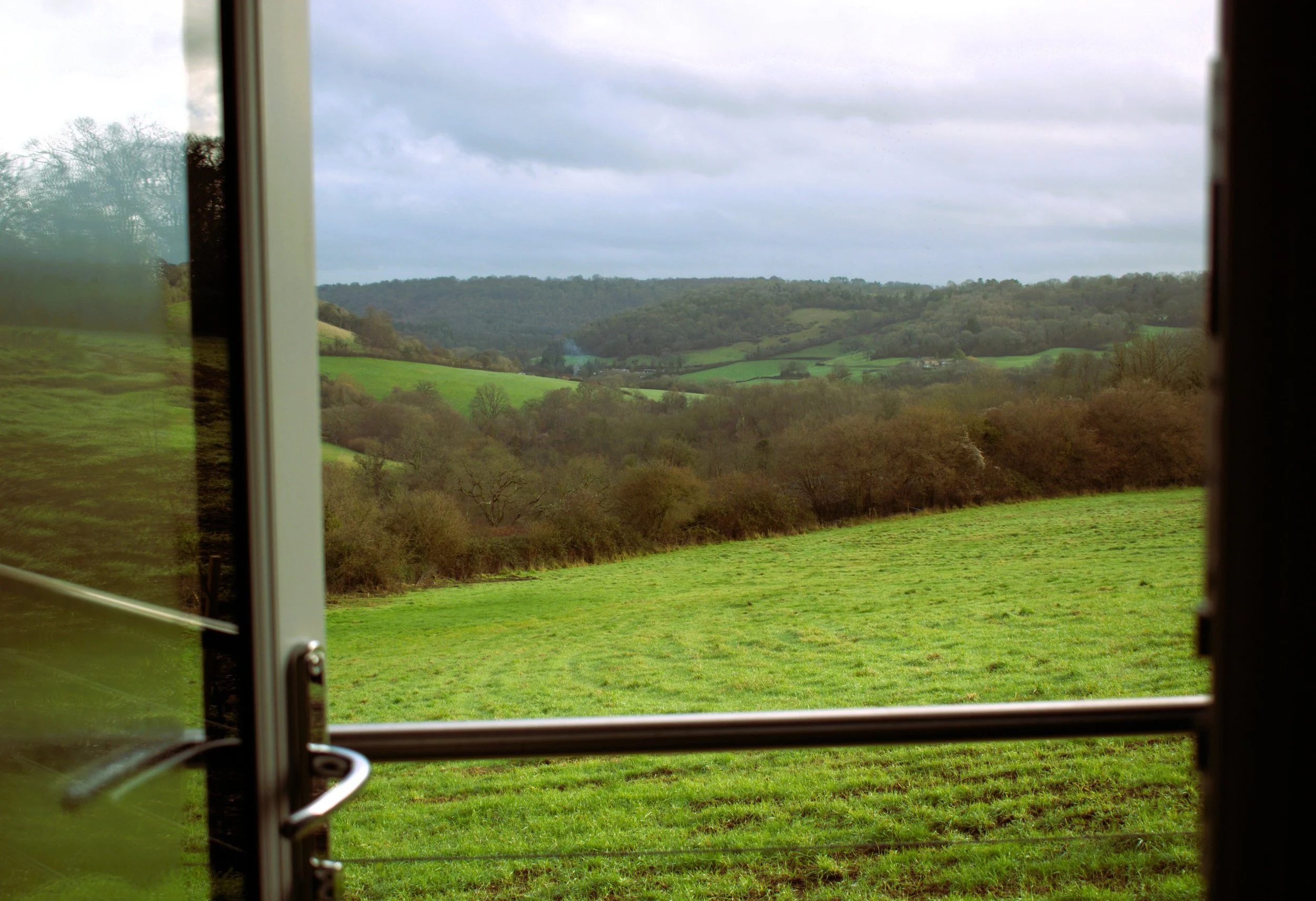 View of green rolling hills and trees through an open door to a cabin balcony on a cloudy day.