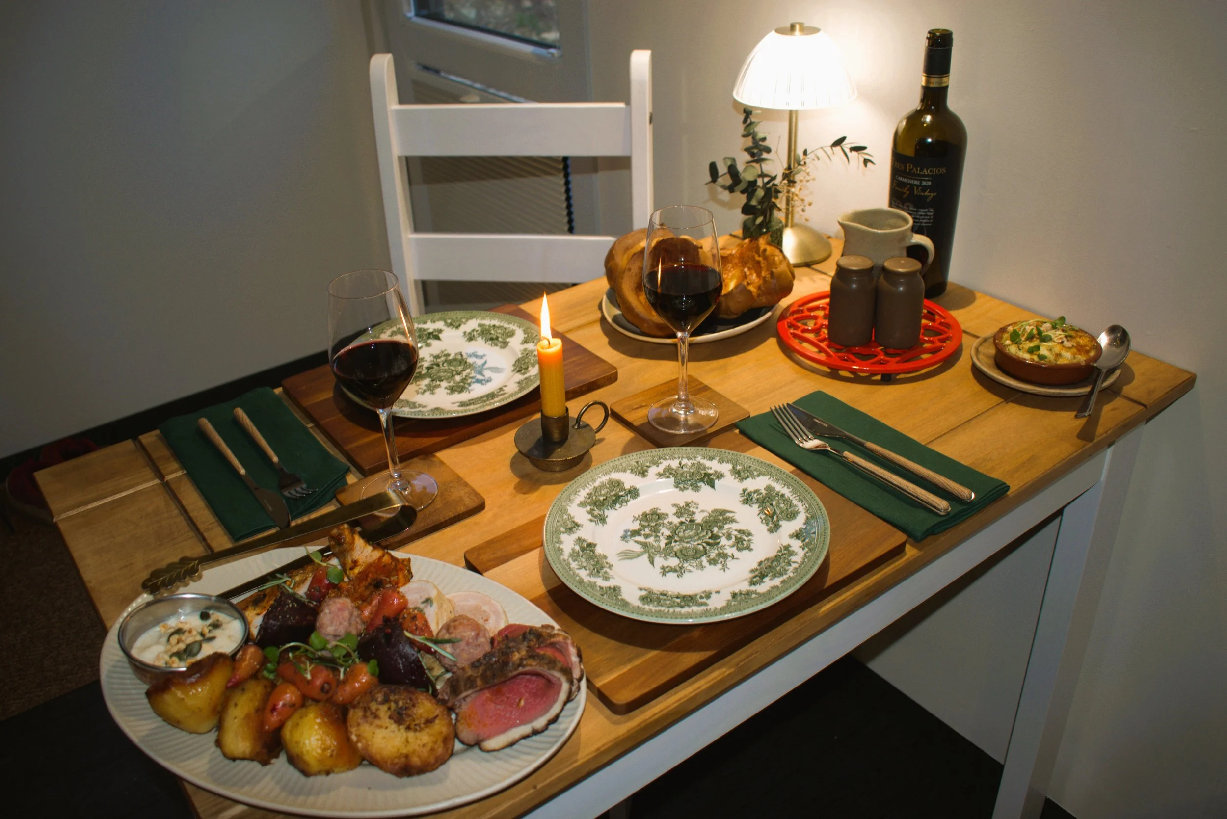 A dining table set with a Sunday Roast dinner, a glass of red wine, cutlery, a candle, a bottle of wine, with a white chair, a small lamp, and decorative items in the background.